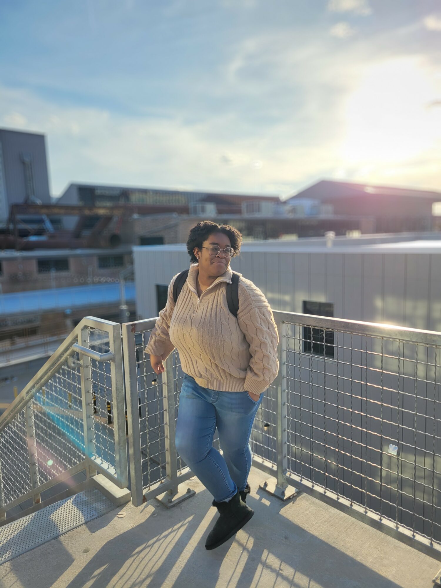 Person with glasses and curly hair standing on a balcony with a cityscape background, sunlight shining, wearing a beige sweater and jeans.