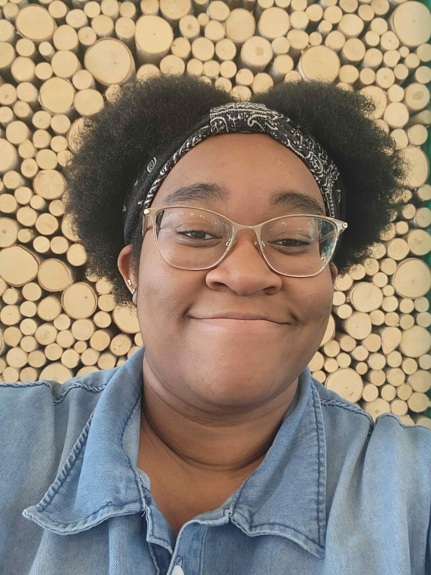 Person with glasses and headband lying on a background of stacked wood logs, wearing a denim shirt.