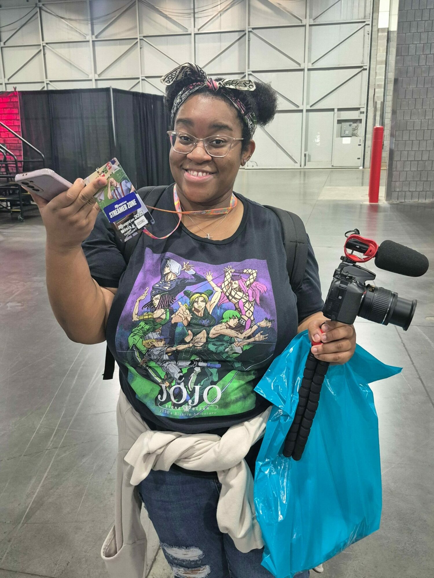 Person smiling, holding a camera and a small item, wearing a graphic t-shirt, glasses, and a blue bag, standing indoors.