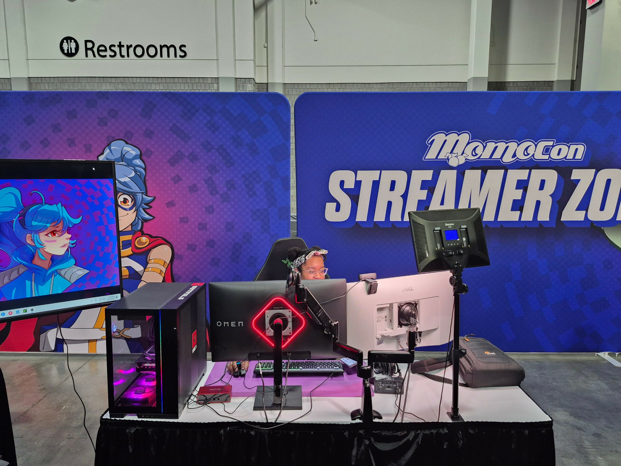 Two people sit at a gaming station with monitors and gaming equipment, in front of a blue backdrop with 'Momocon STREAMER ZONE' text.