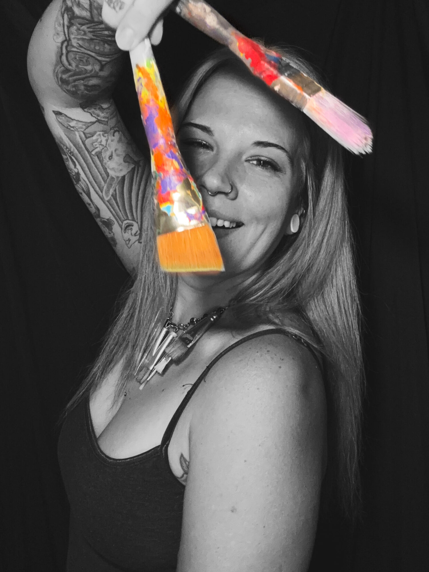 Woman smiling, holding paintbrushes with colorful paint, black and white background, tattoos visible on arm, wearing a tank top.