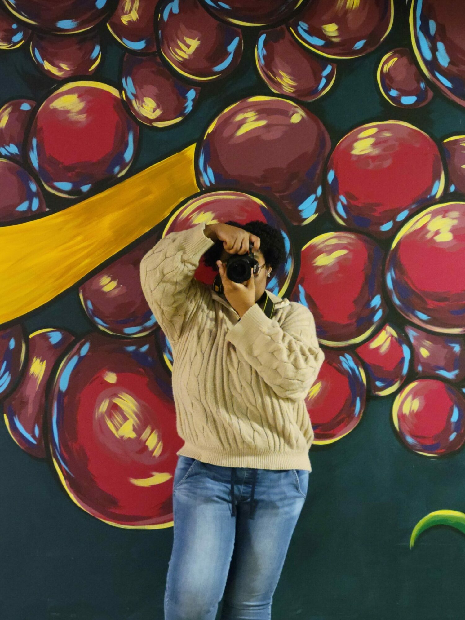 Person taking a photo with a camera in front of a colorful mural with red and yellow abstract shapes.
