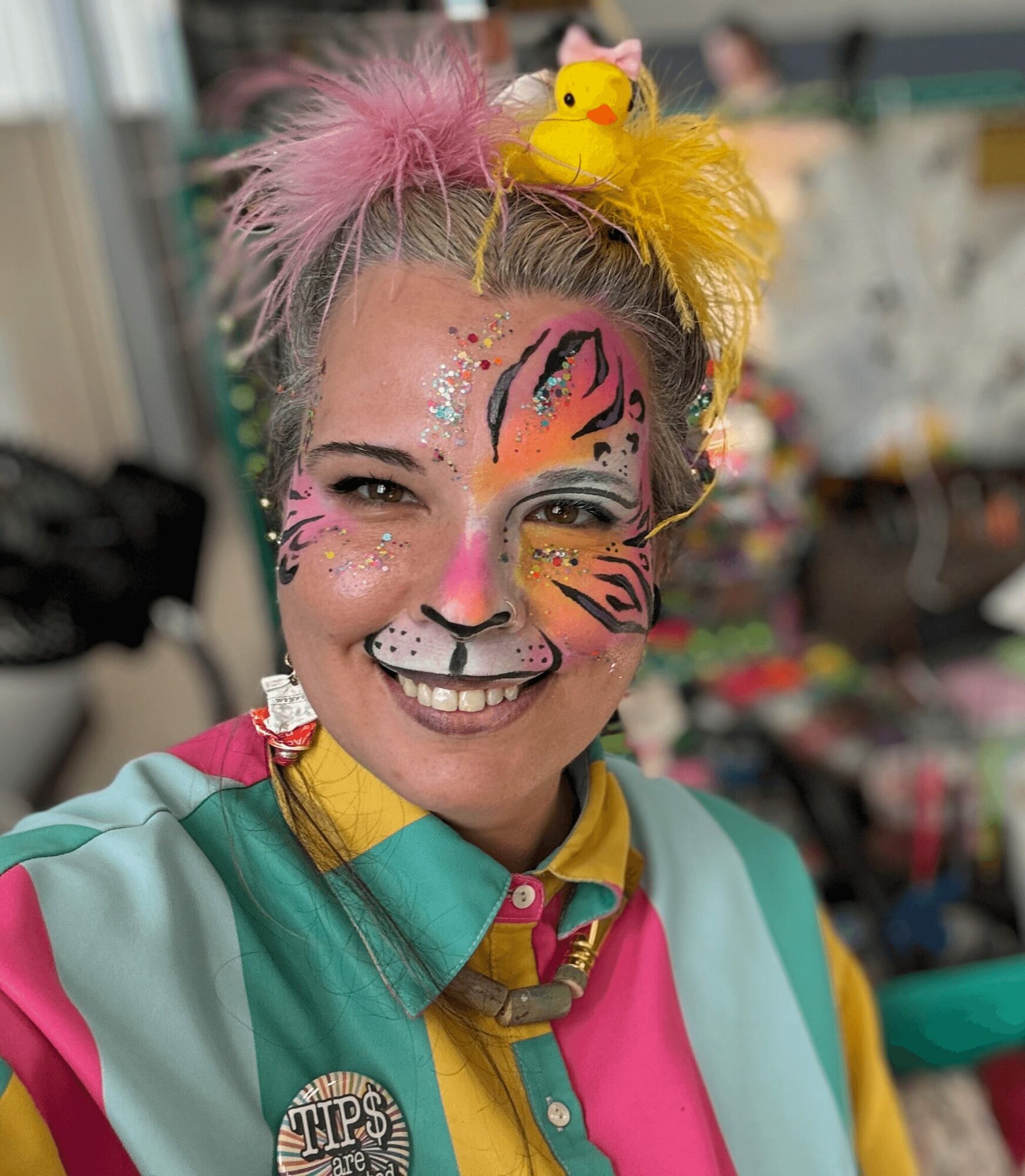 Person with colorful face paint, pink and yellow hair accessories, and a bright, multicolored outfit smiling.