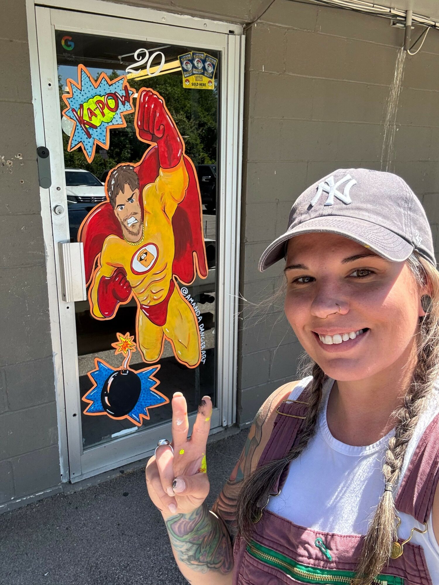 Person with long hair, wearing a white cap and pink jacket, smiling and making a peace sign next to a colorful cartoon cutout.