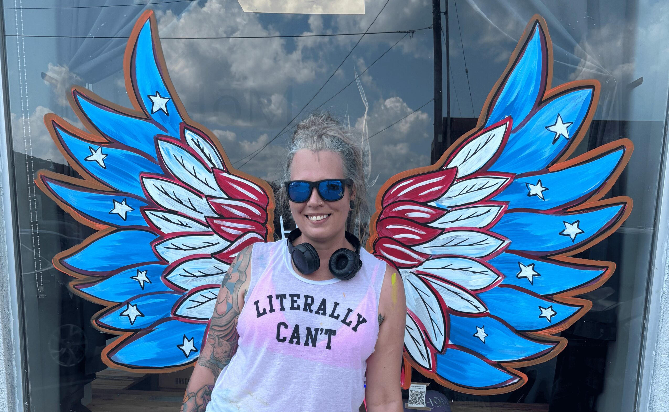 Woman with sunglasses and headphones, smiling, standing in front of colorful wing art, wearing a white shirt with text.