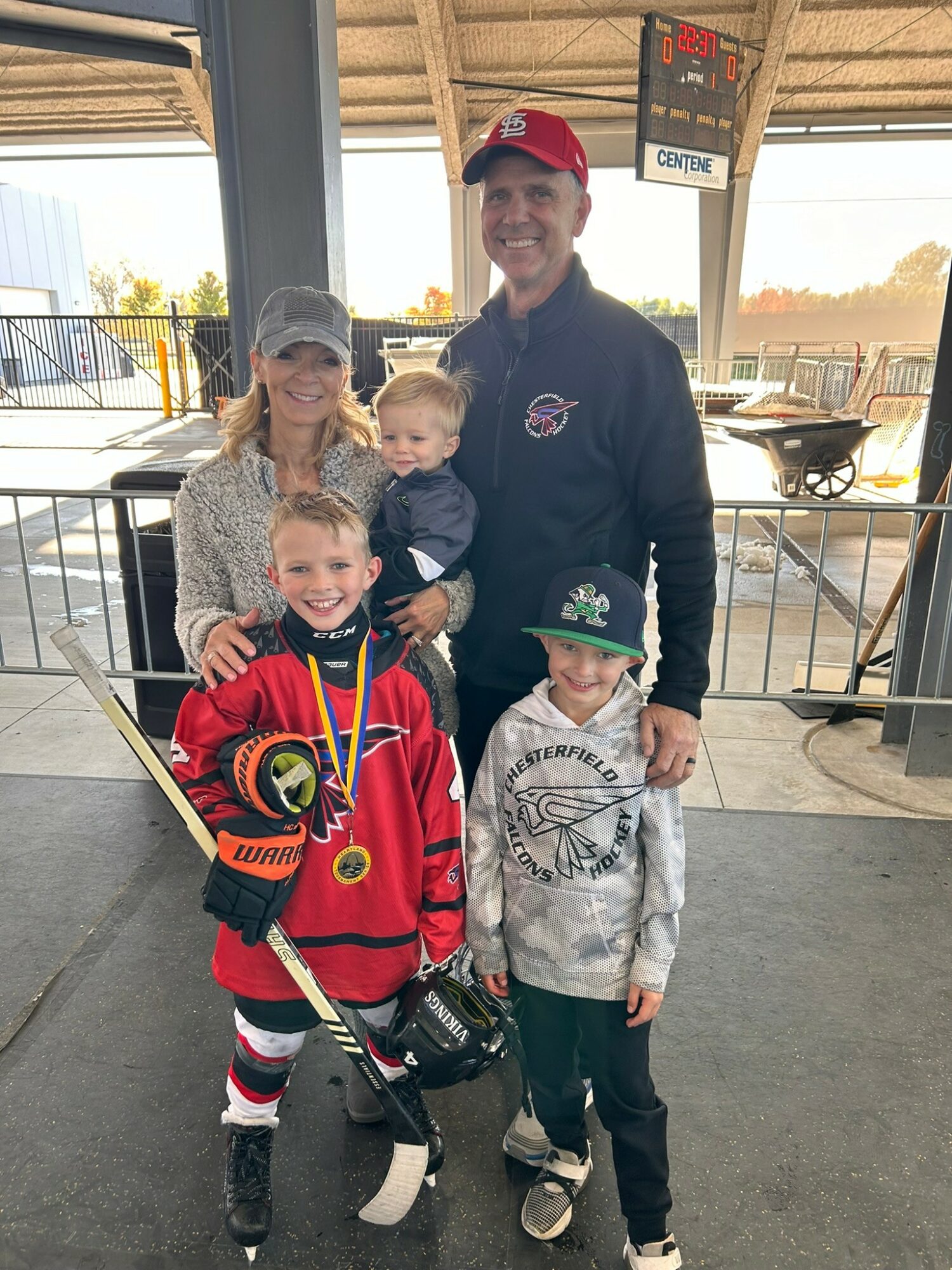 Group of five people, including children, standing outdoors with a man in a black jacket and red cap, smiling.