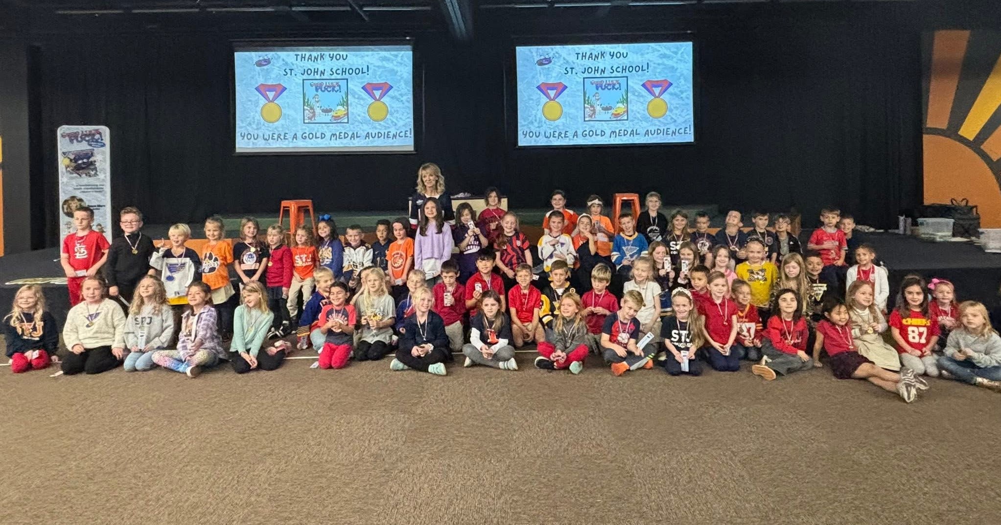 Large group of children and adults posing indoors in front of two screens displaying colorful graphics.