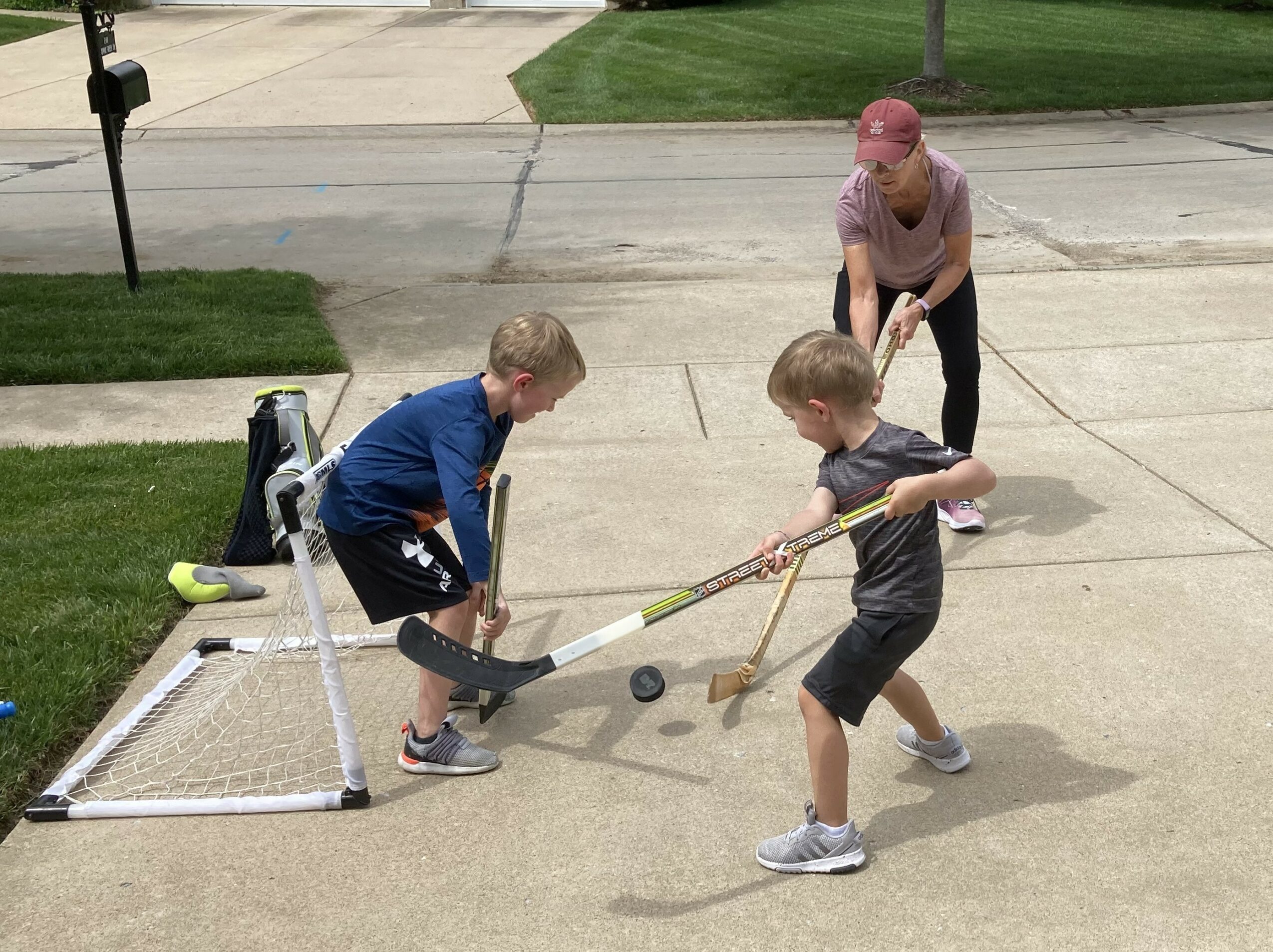 Two children play street hockey with a woman watching on a sidewalk, a small goal, and a ball present.