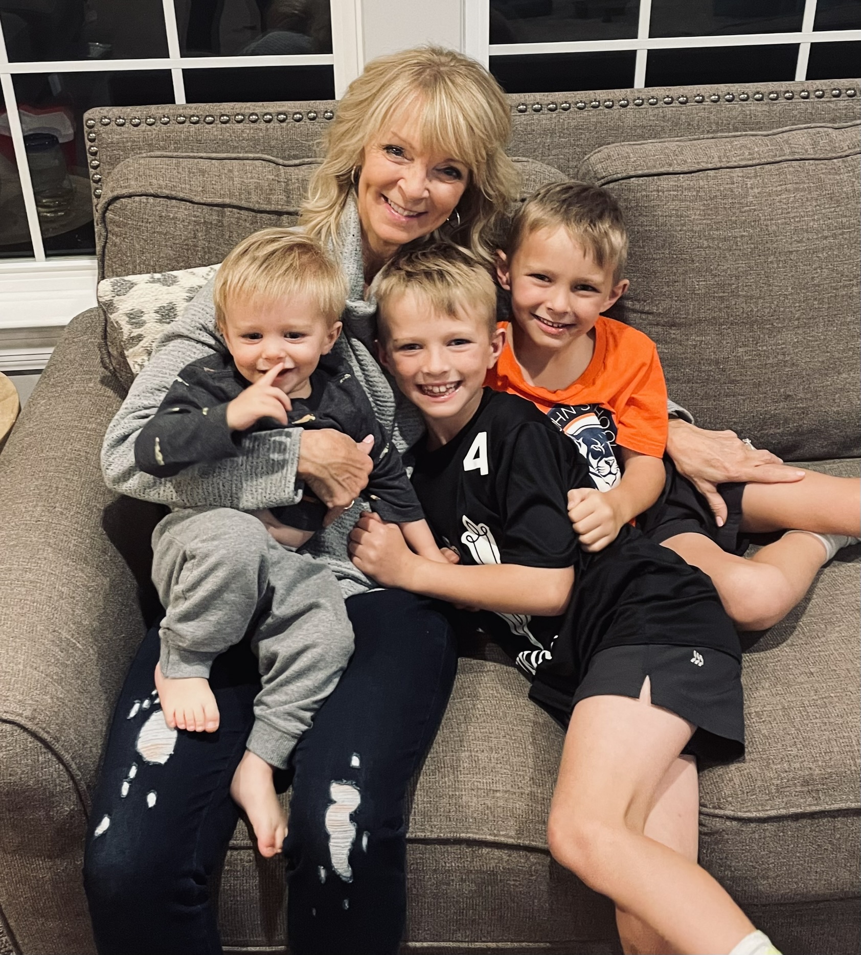 A woman with three children sitting on a sofa, smiling, in a living room with windows behind them.