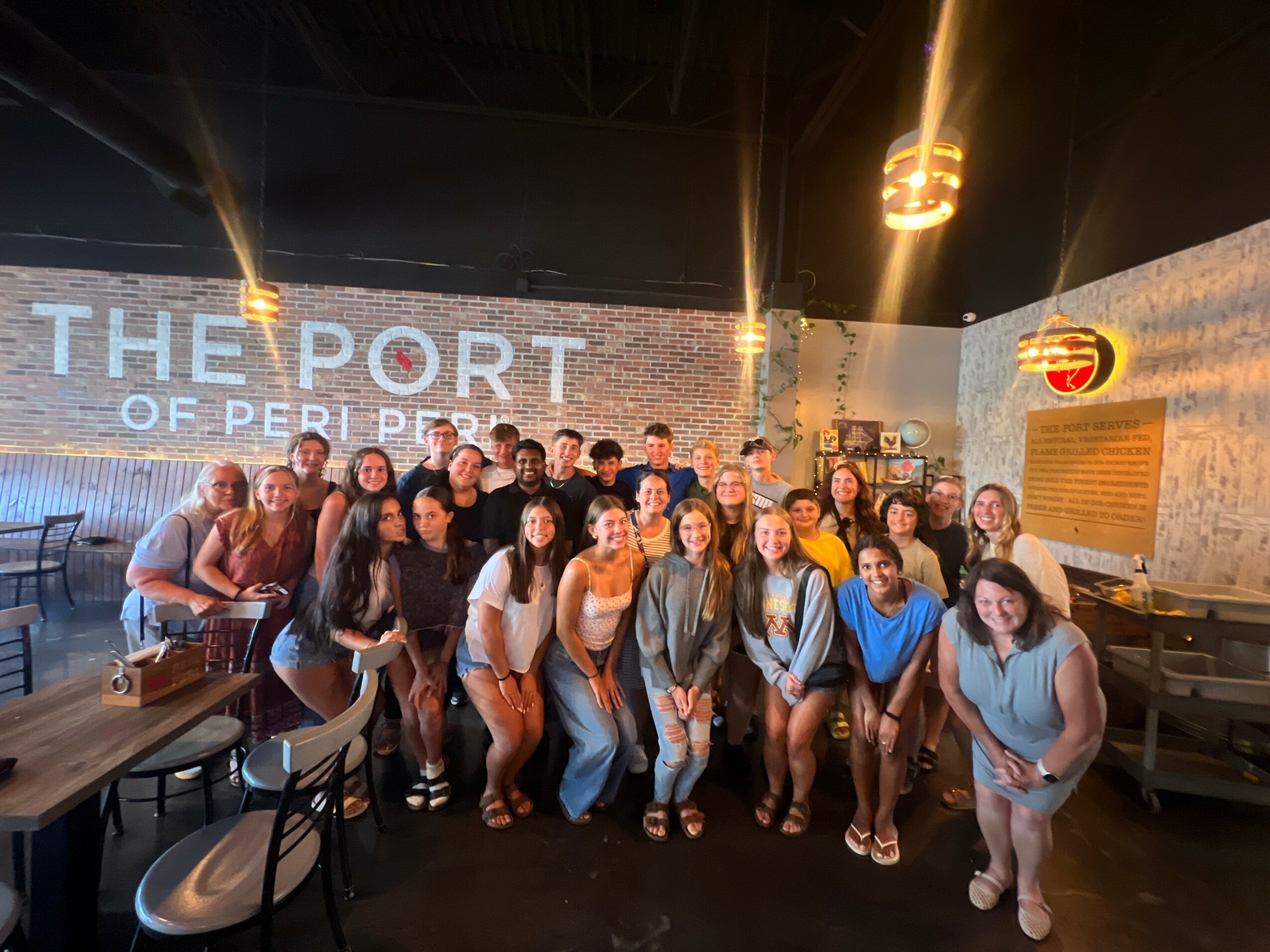 Group of people posing in a restaurant with brick walls and a large illuminated sign reading 'THE PORT OF PERI PERI'.