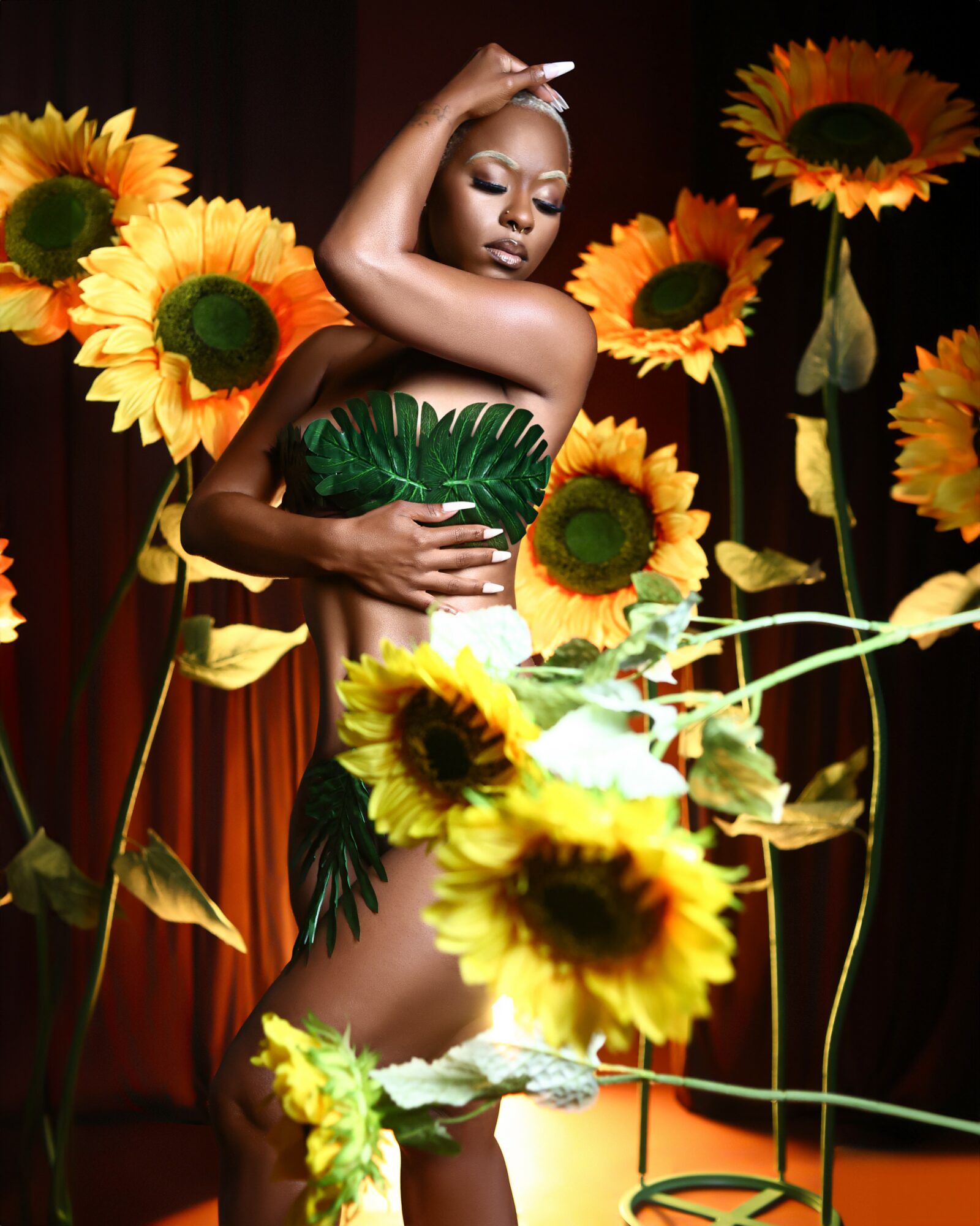 A woman in a green leaf-themed outfit surrounded by large sunflowers, with her eyes closed and hand on her head.