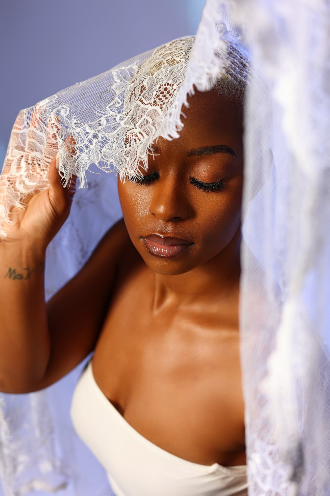 Woman with closed eyes holding lace fabric near her face, partially covered by lace, against a blue background.