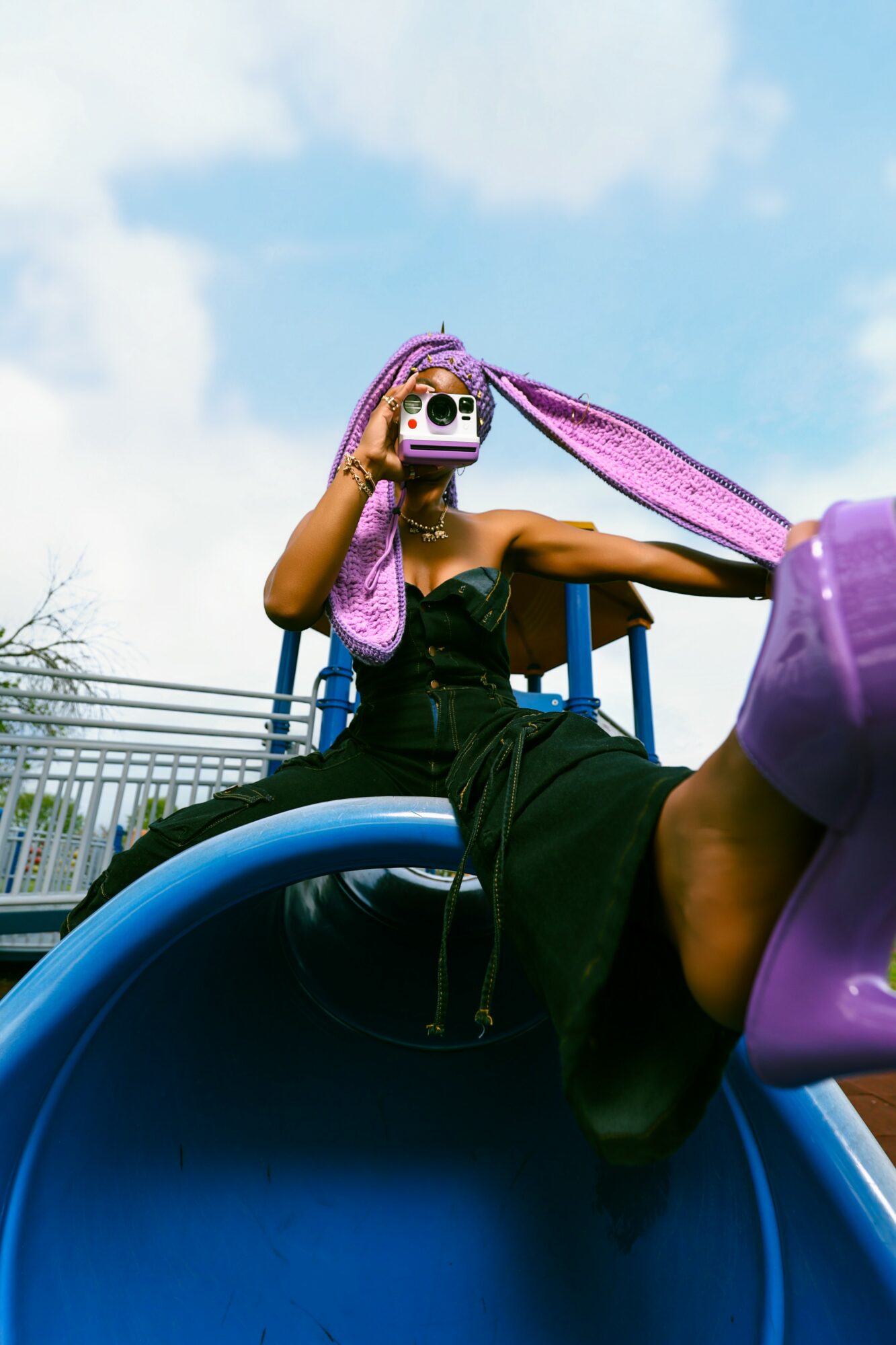 Person sitting on a slide holding a camera, wearing a purple headscarf and black dress, with a blue sky background.