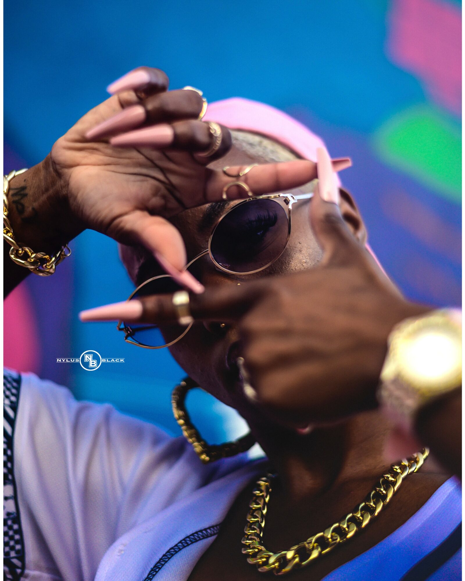 Person wearing sunglasses, gold chain, and jewelry, adjusting head with hands, colorful background, close-up shot.