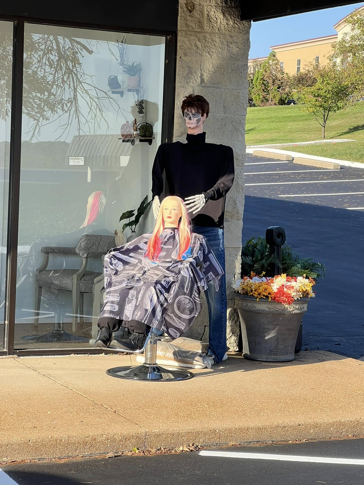 Person standing outside a glass storefront with a mannequin head on a chair, next to a flower pot, on a sunny day.