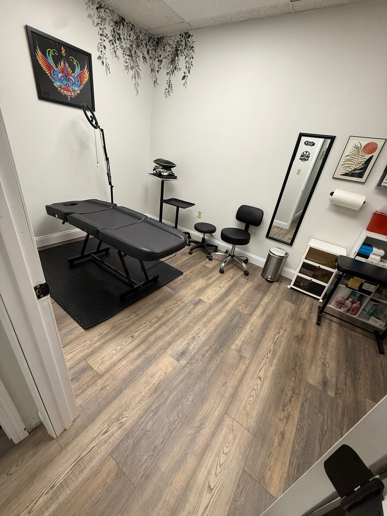 Room with massage table, stool, mirror, wall art, and storage shelves, with wooden floor and white walls.