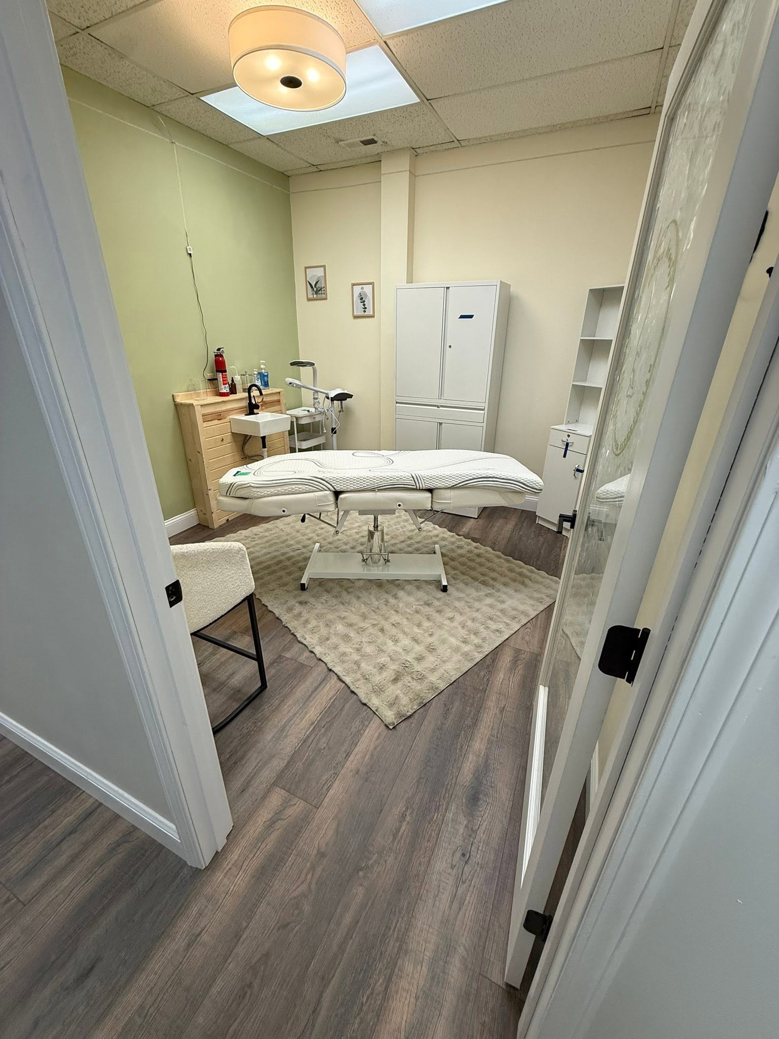 Medical examination room with bed, white cabinet, shelves, and medical supplies, viewed from doorway.