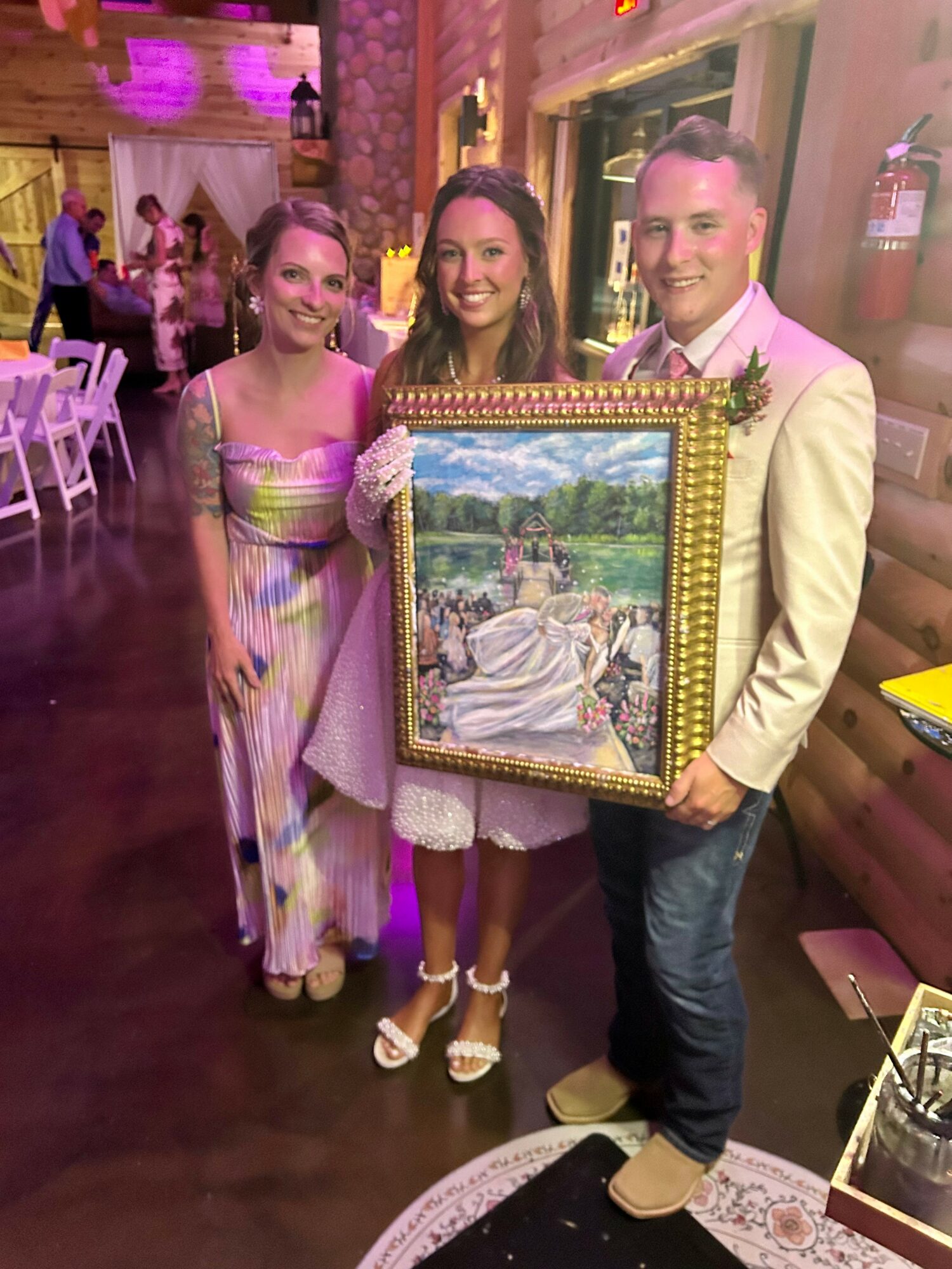 Three people standing indoors, holding a framed painting, smiling, with a decorated background and other guests in the distance.