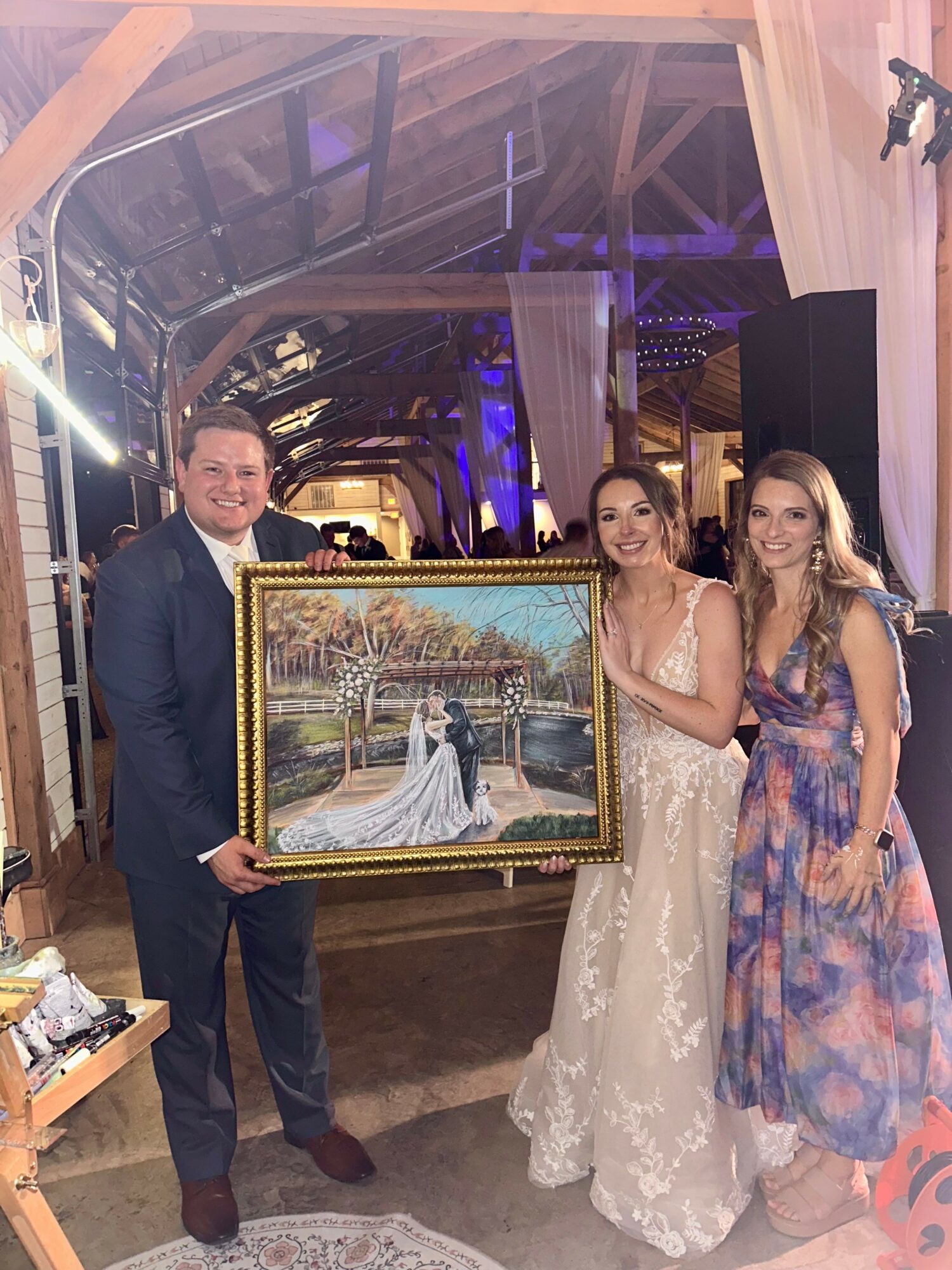 Three people holding a framed wedding photo, standing indoors with wooden beams and purple lighting, smiling.