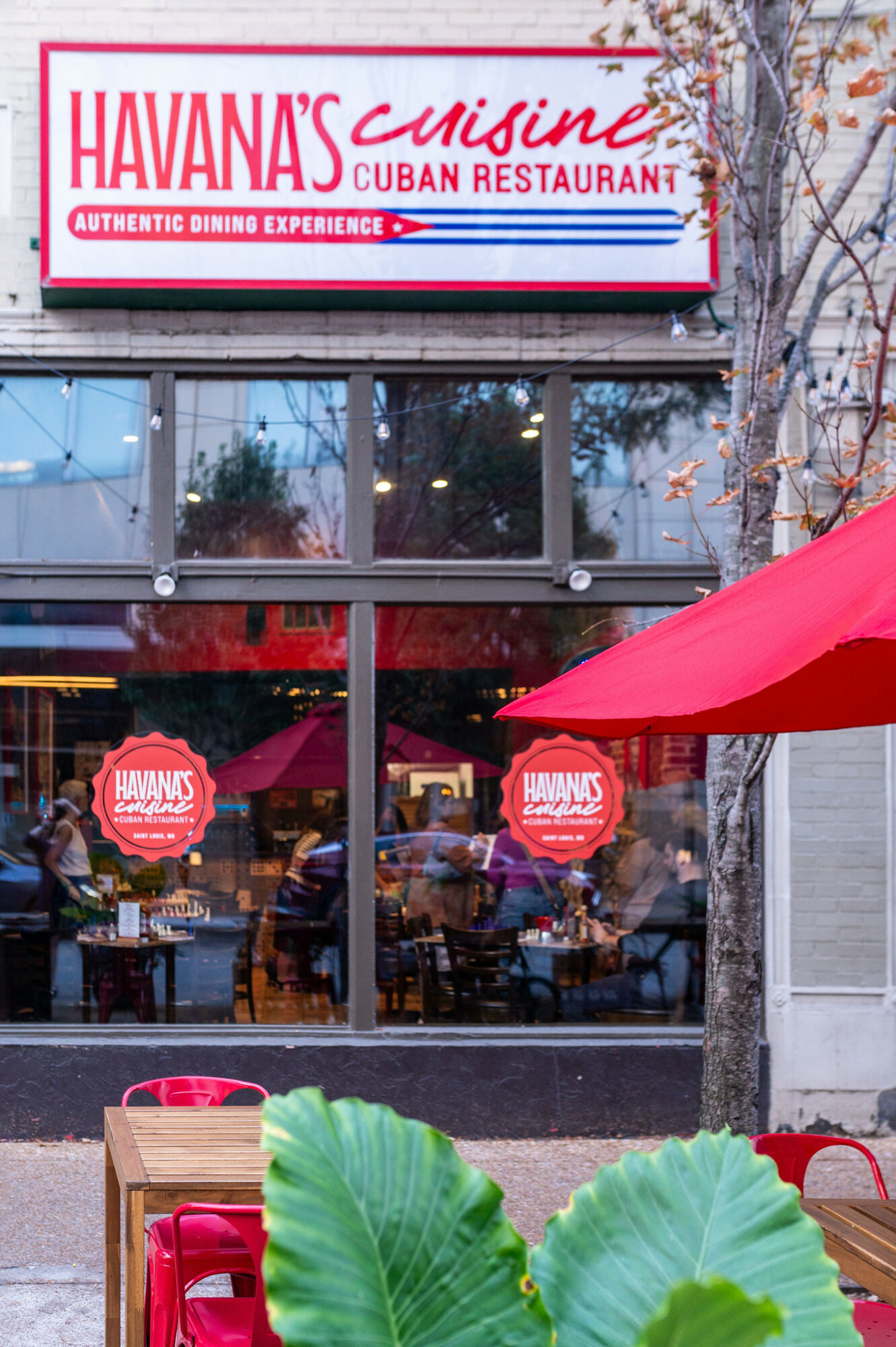 Restaurant storefront with large glass windows, red umbrellas, and outdoor seating, with a sign reading 'Havana's Cuisine Cuban Restaurant'.