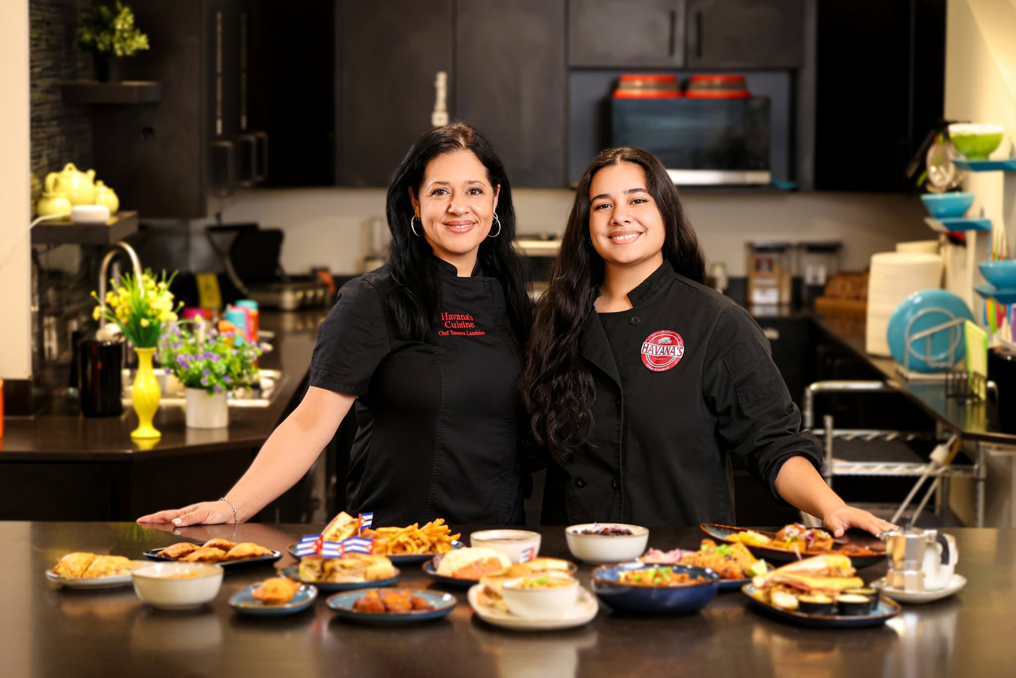 Two women in black shirts stand behind a table with various baked goods in a kitchen, smiling at the camera.