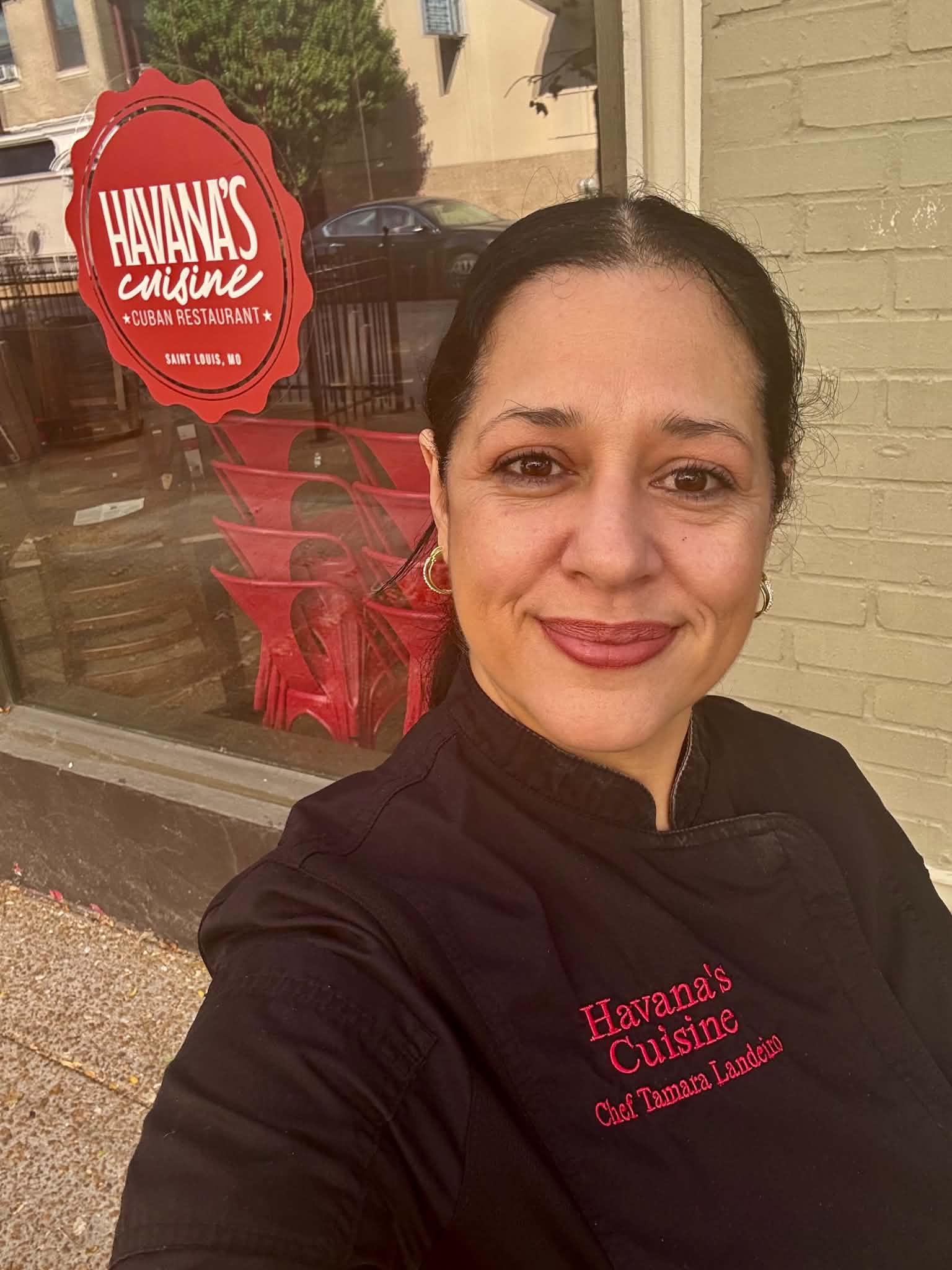 Woman taking a selfie outside a restaurant with a red sign, wearing a black jacket with red text, smiling.