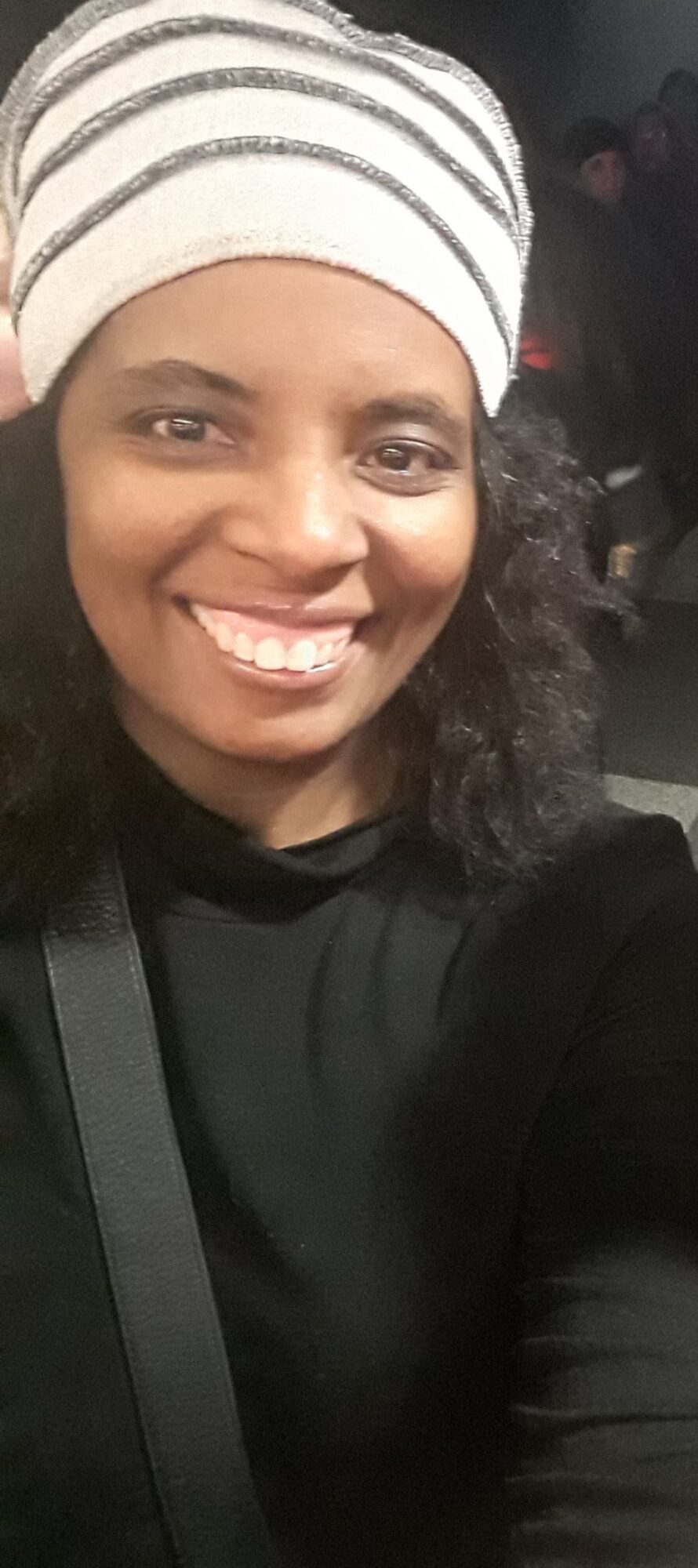 Smiling woman wearing a white striped beanie and black top, with curly hair, in a close-up shot.