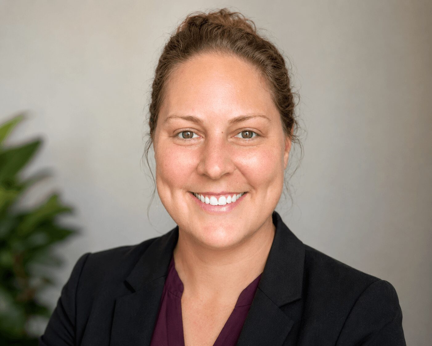 Portrait of a smiling woman with brown hair in a bun, wearing a black blazer and purple top, against a neutral background.