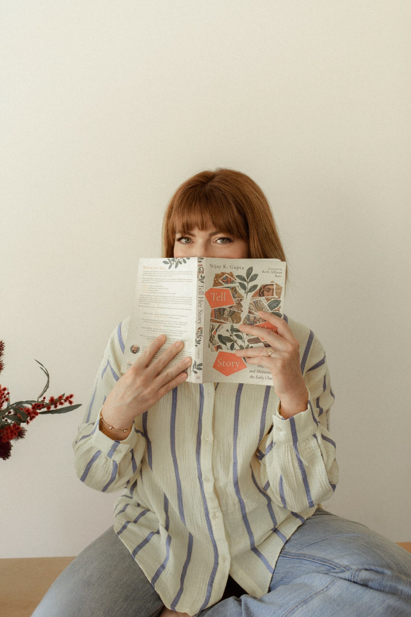 Person with reddish hair reading a book, sitting against a plain white wall, wearing a striped shirt.