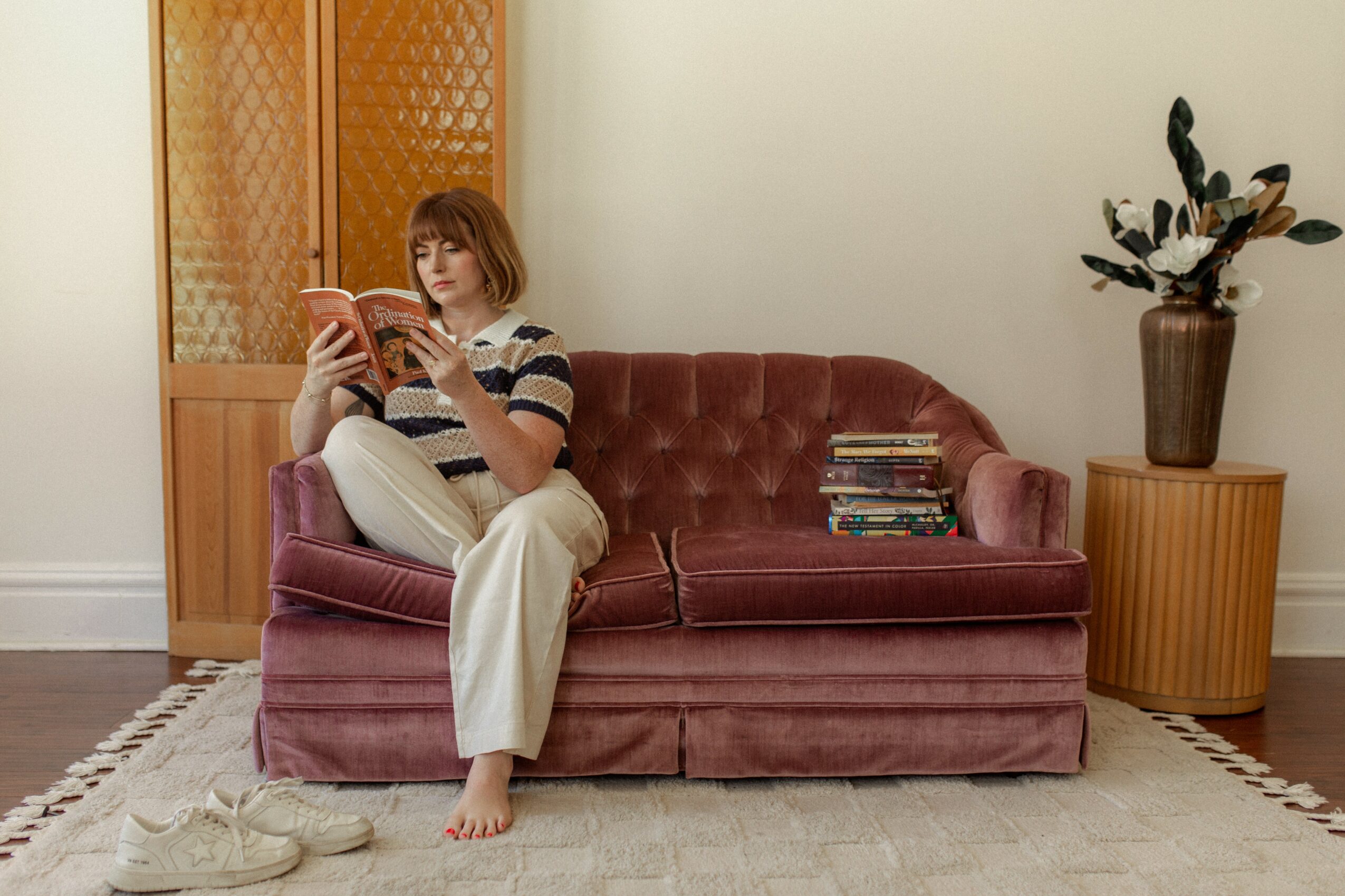 Woman sitting on a pink velvet sofa reading a book, with a stack of books beside her, in a living room.