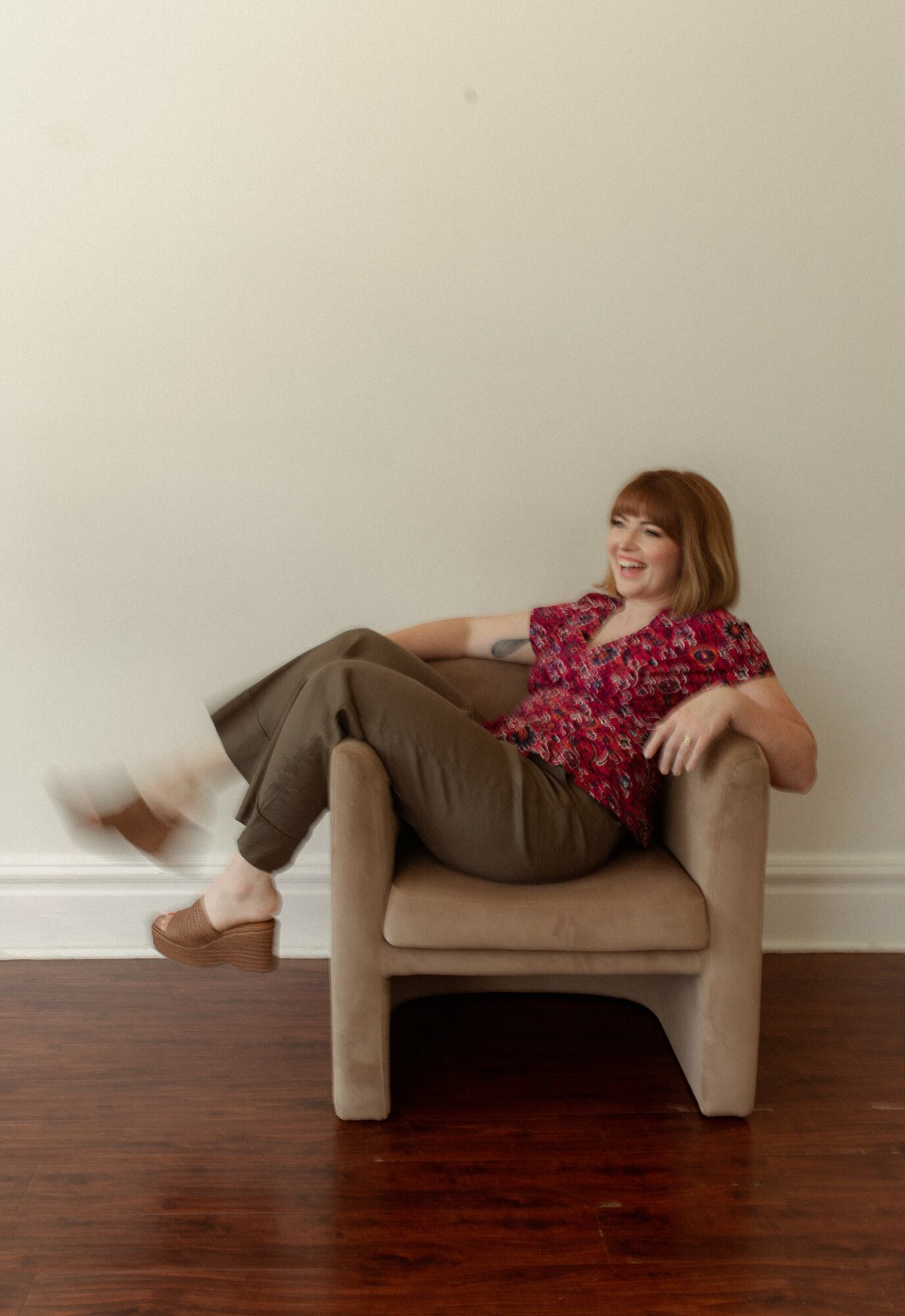 Woman sitting on a beige armchair with a plain white wall background, smiling, wearing a red floral blouse and brown pants.