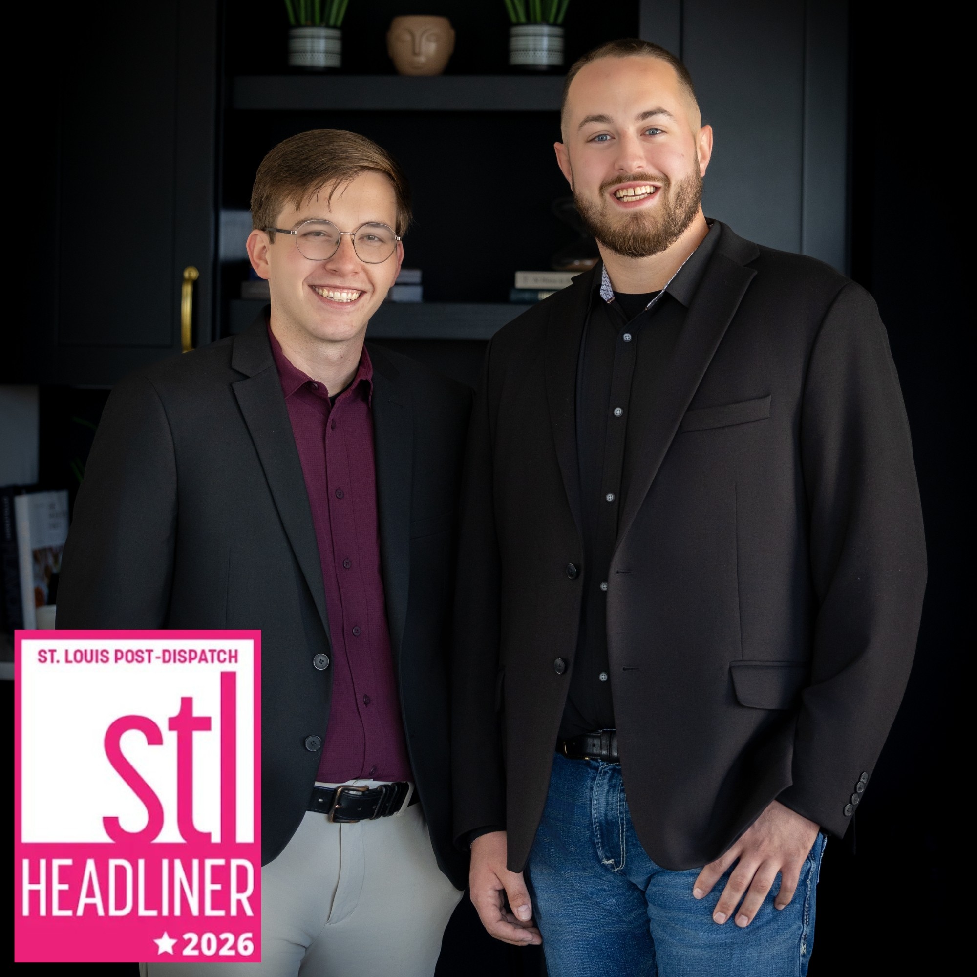 Two men in suits smiling indoors with dark background and shelves, one with glasses, both with short hair and beards.