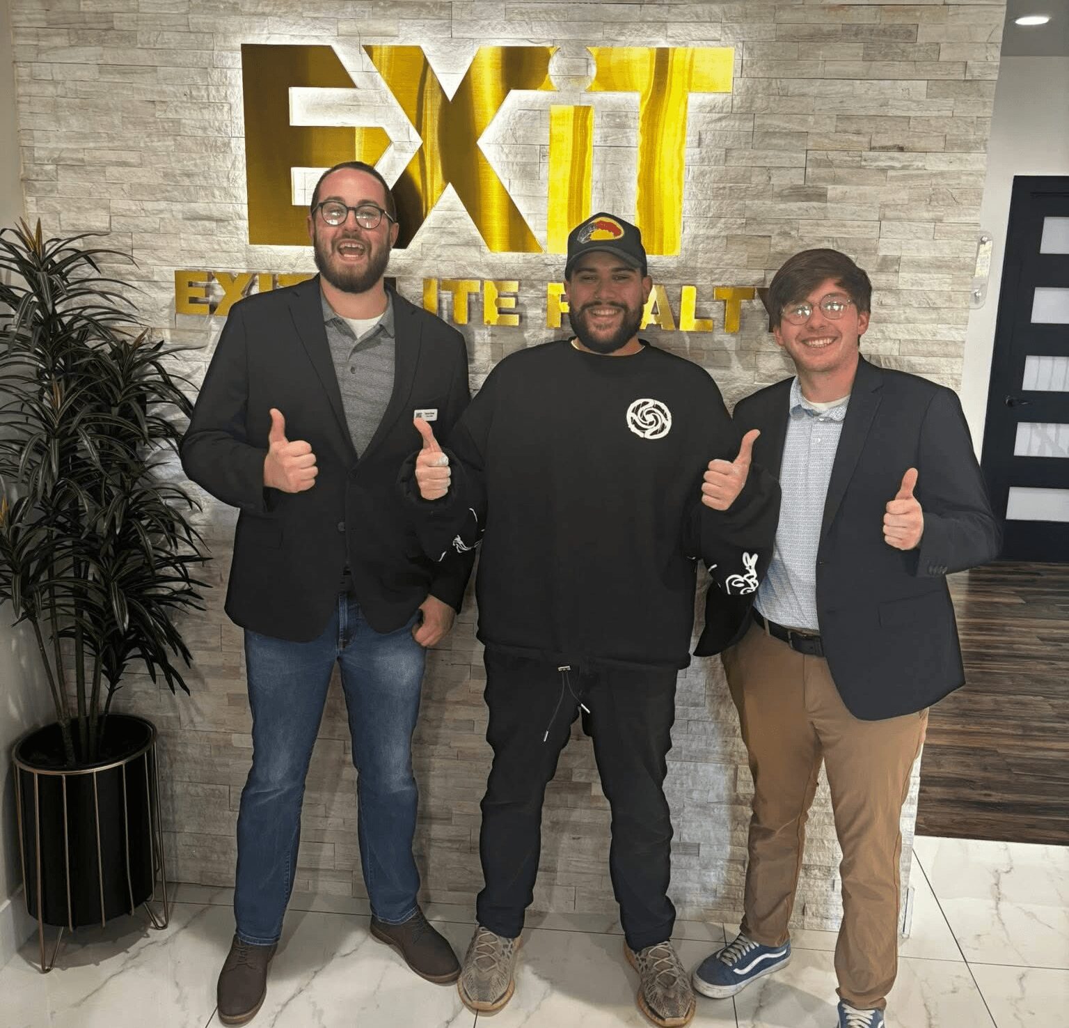Three men standing in front of a wall with a large gold and white EXIT sign, smiling and giving thumbs up.