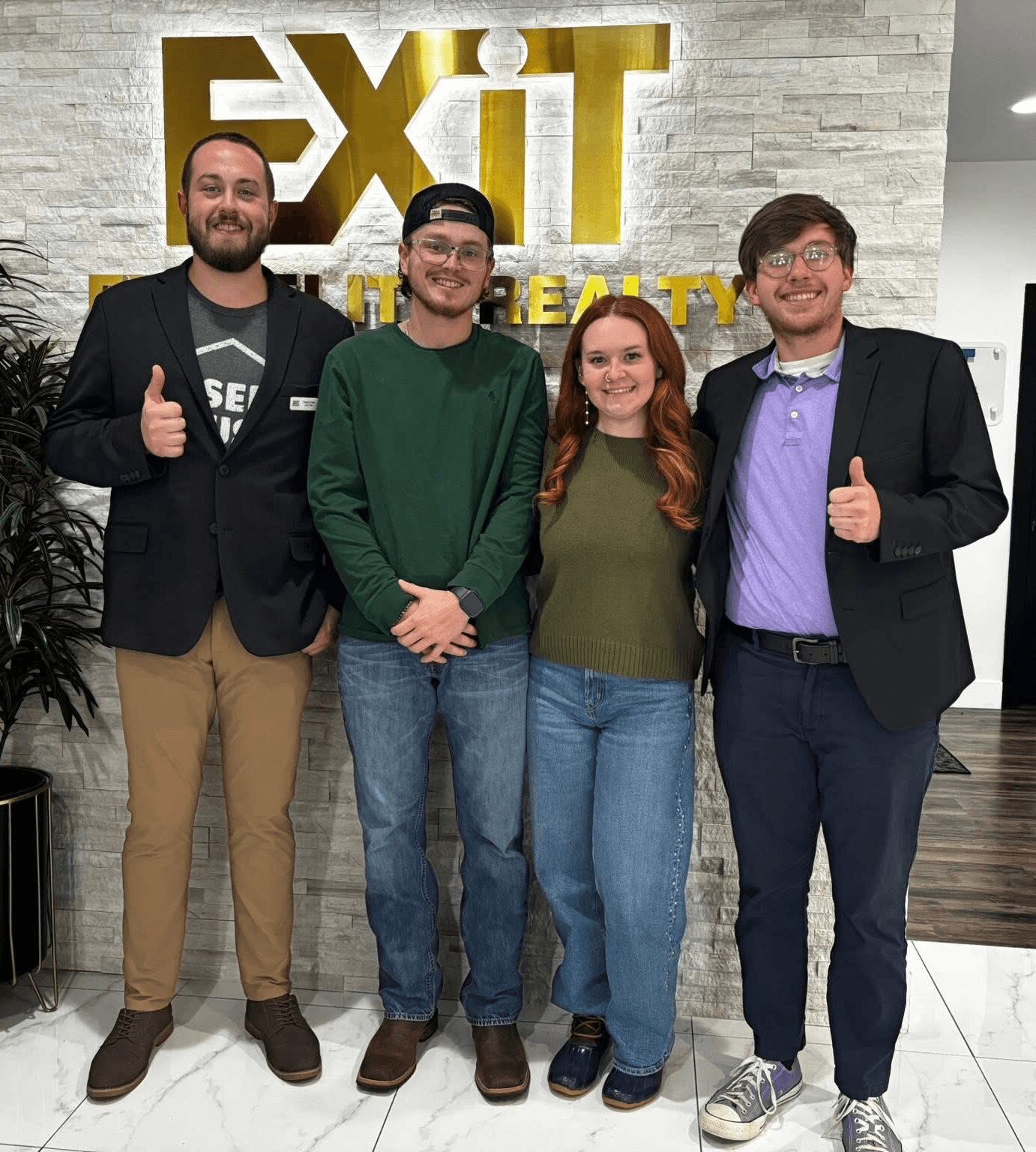 Four people standing indoors in front of a wall with a large illuminated sign reading 'EXIT'. They are smiling, with two giving thumbs-up gestures.