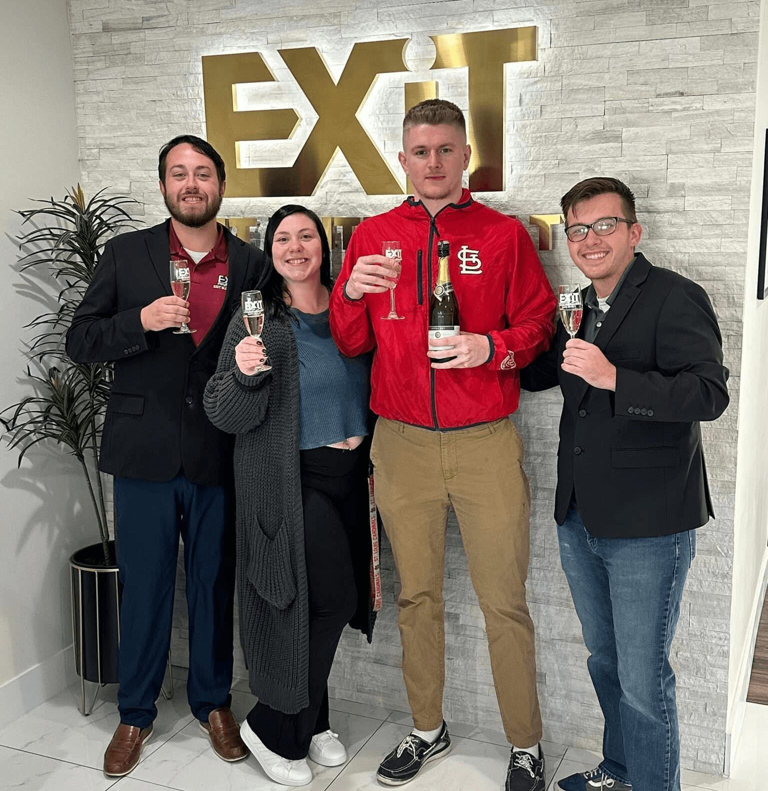 Four people holding glasses, standing in front of a wall with a large gold 'EXIT' sign, smiling at the camera.