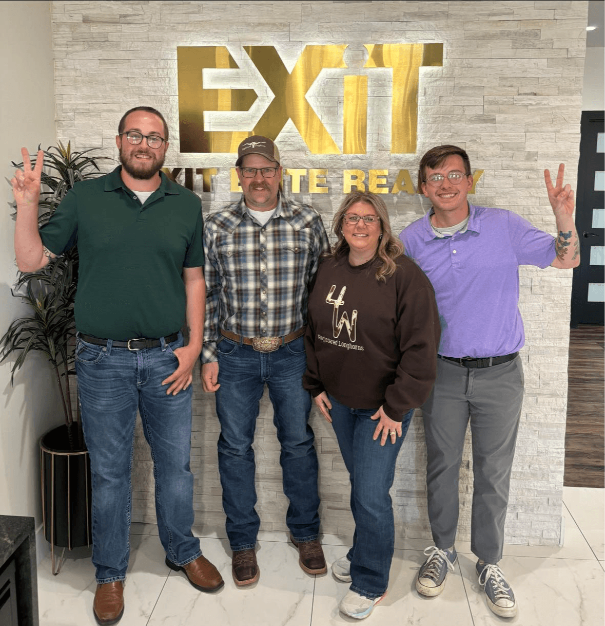 Four people standing in front of a wall with a large gold and white sign reading 'EXIT'. They are smiling and making peace signs.