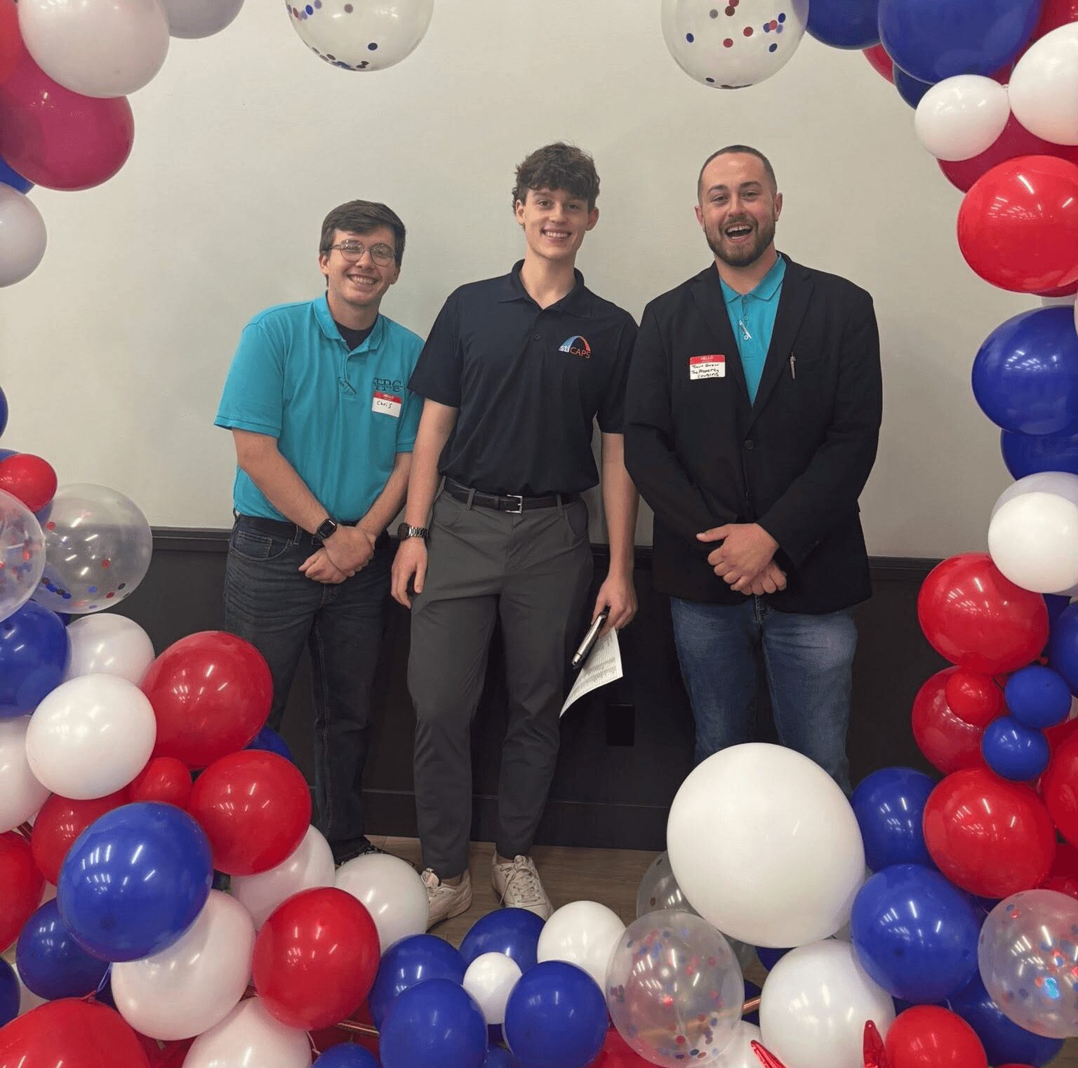 Three people stand inside a balloon arch with red, white, and blue balloons, smiling at the camera.