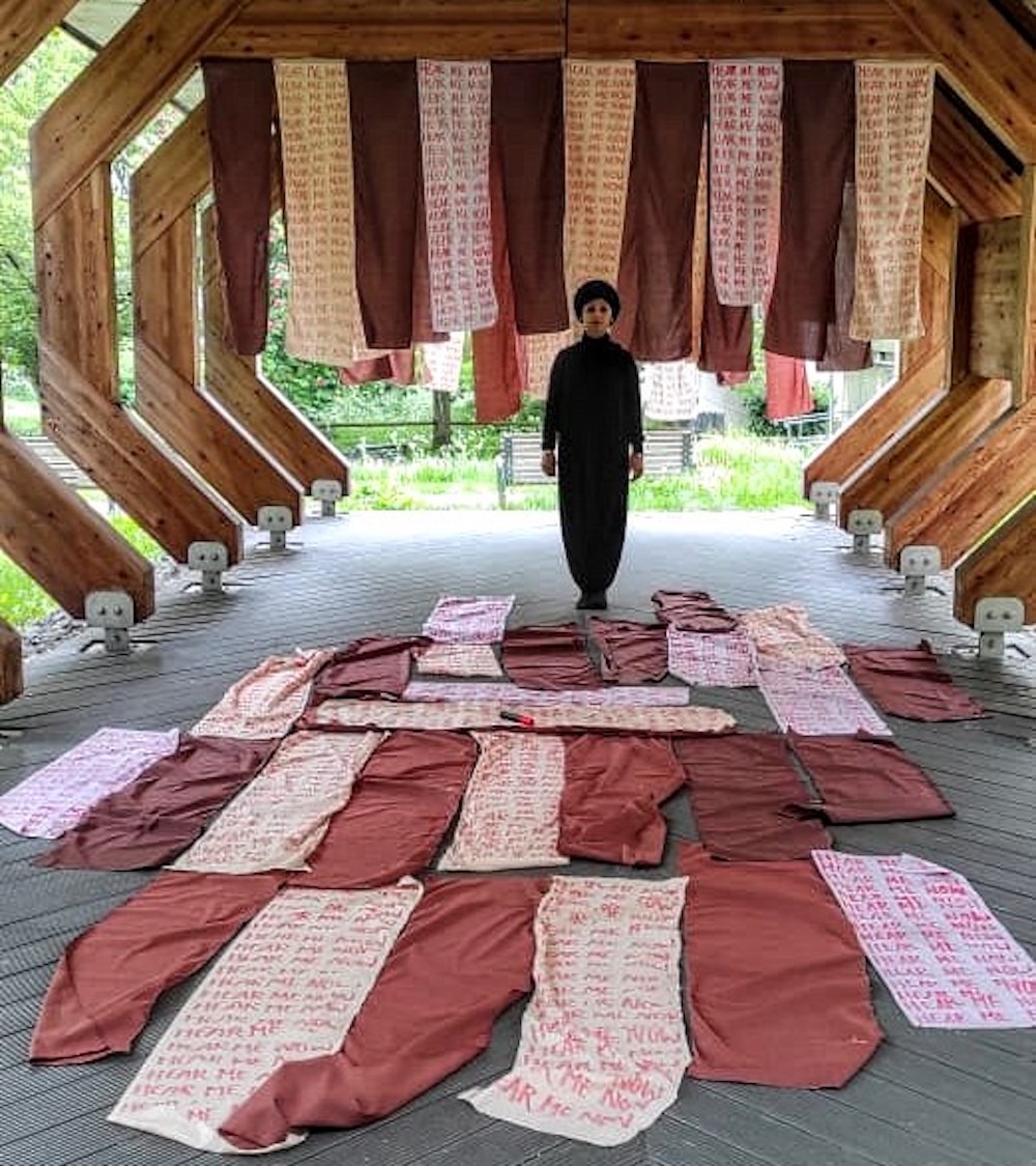 Person standing under a wooden structure with hanging fabric and cloths spread on the floor, greenery visible outside.