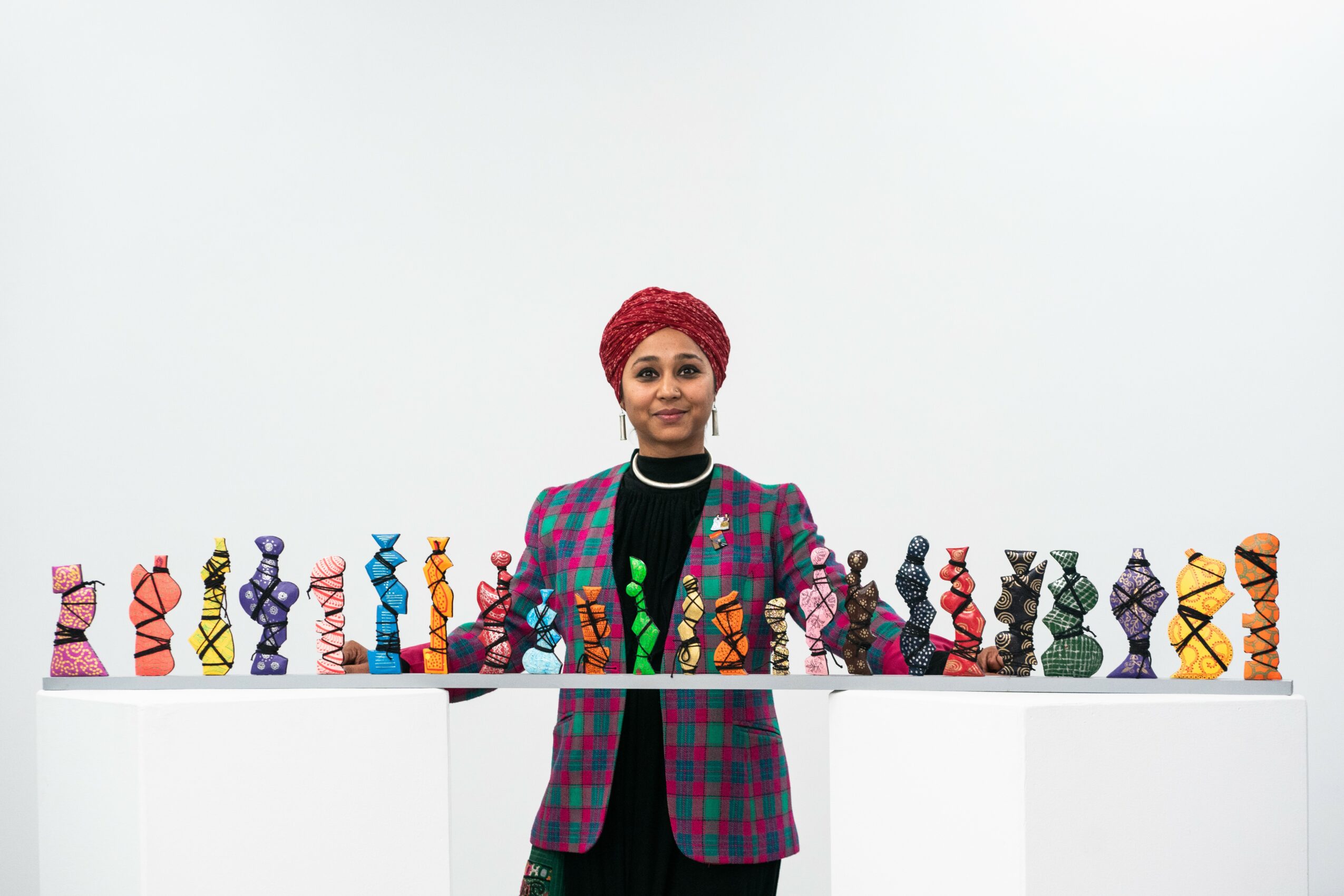 Woman with red headwrap and colorful blazer stands behind a table with various colorful sculptures.