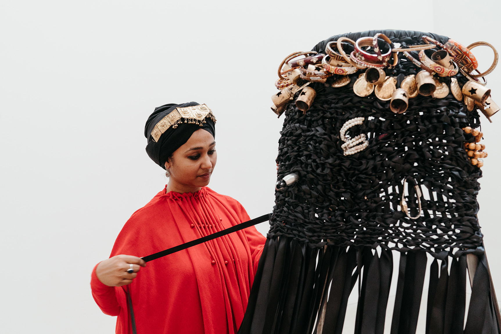 A woman in a red dress and headscarf pulls a large, black, intricately decorated sculpture with a stick.