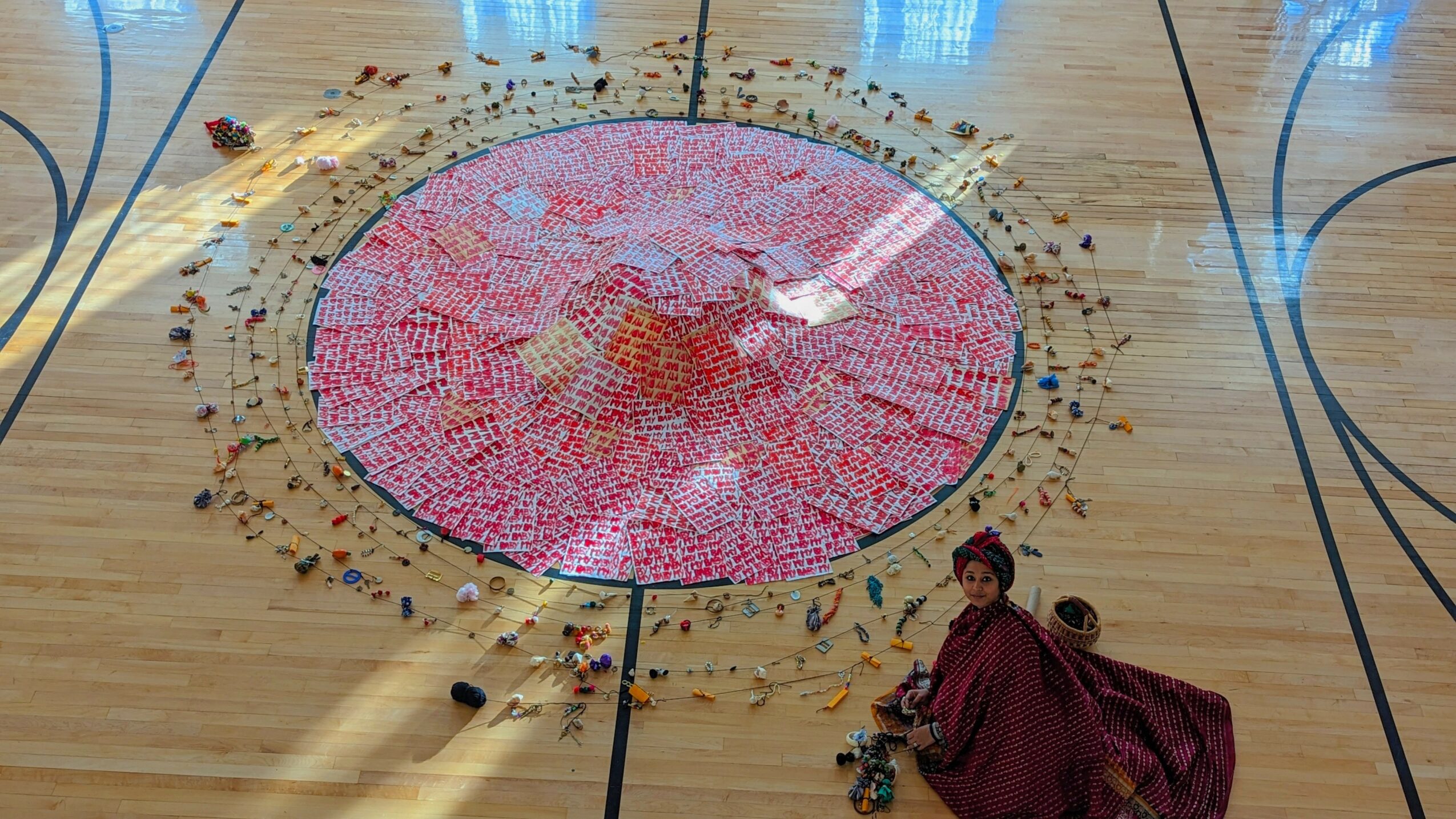 A person in a maroon dress lies on a basketball court near a large, colorful circular mosaic with scattered objects around.