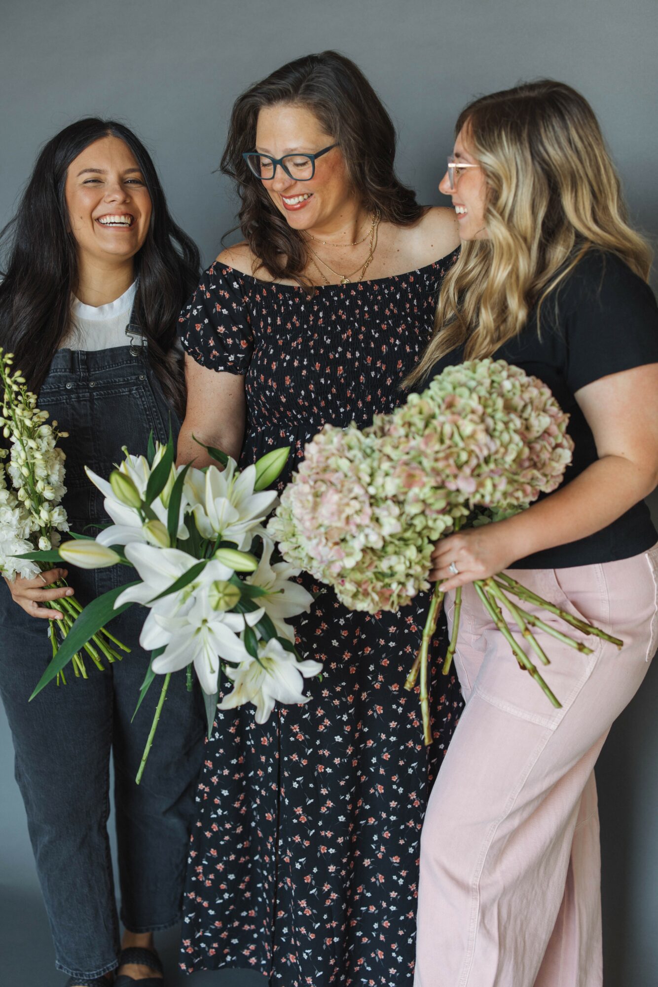 Three women smiling and holding bouquets of flowers, standing together indoors against a plain background.