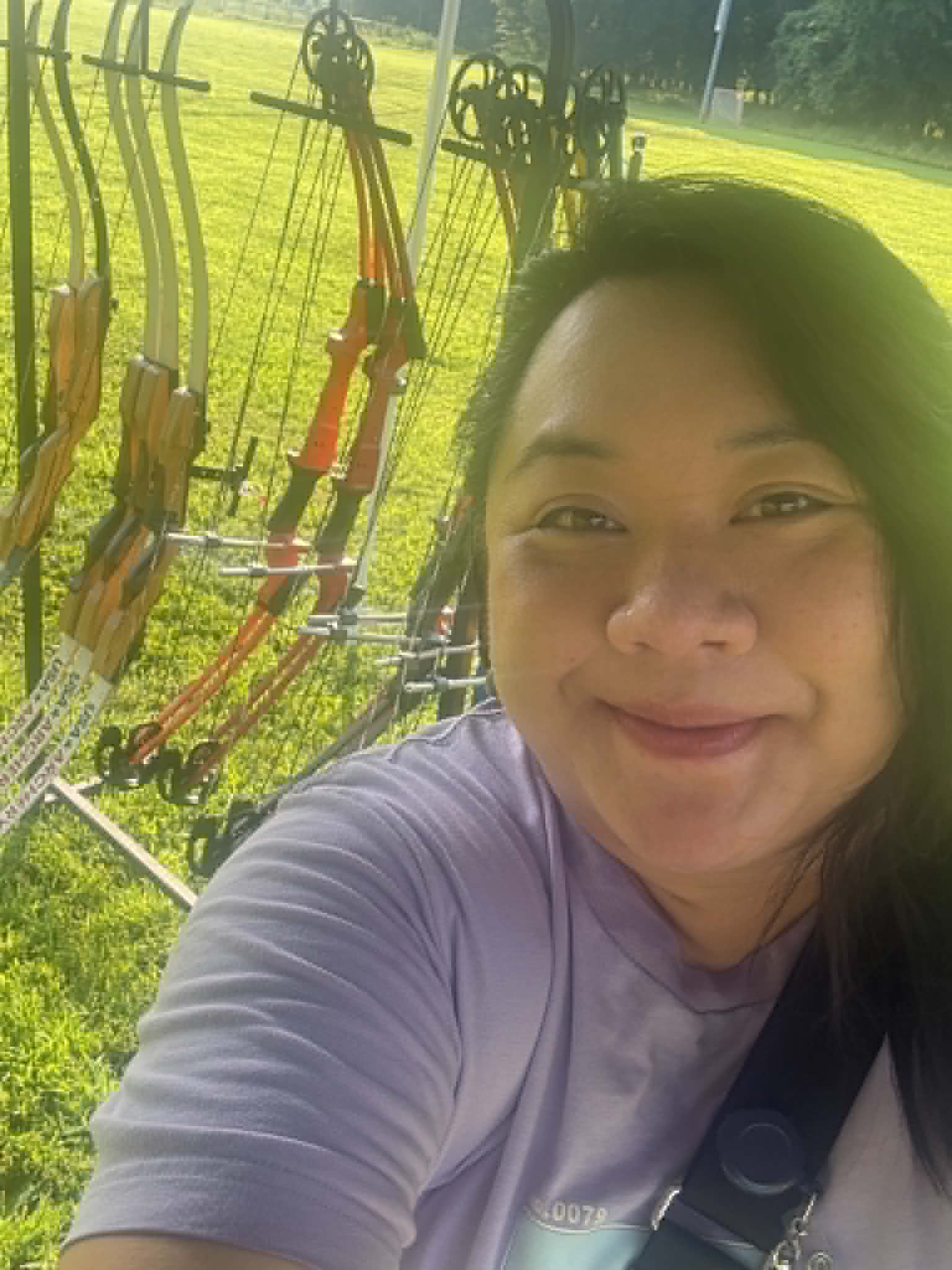 Woman smiling outdoors near archery equipment with bows and arrows, green grass background.