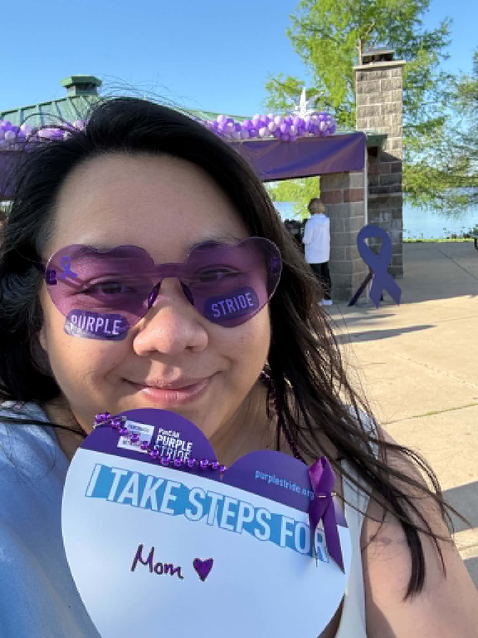 Woman wearing purple sunglasses holding a sign that says 'Take Steps for Mom' outdoors with trees and a stone structure in background.