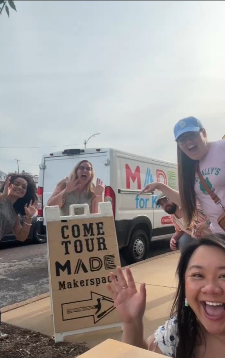 Group of people smiling and waving outdoors near a sign and a van, with a clear sky in the background.