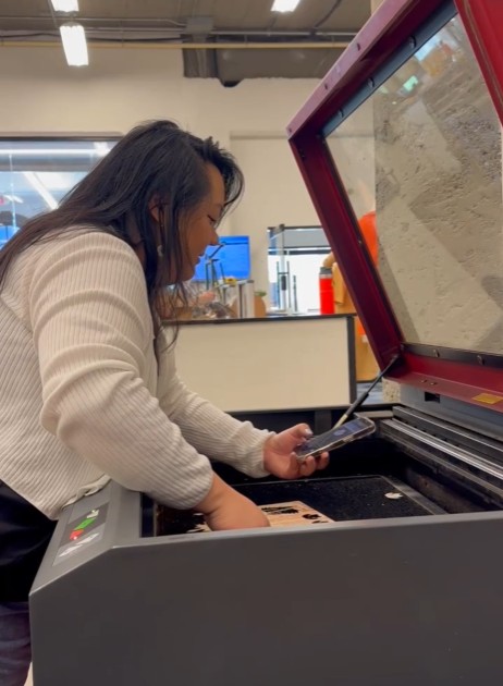 Woman playing piano with a red and black keyboard, looking at a smartphone, in a room with shelves and a window.