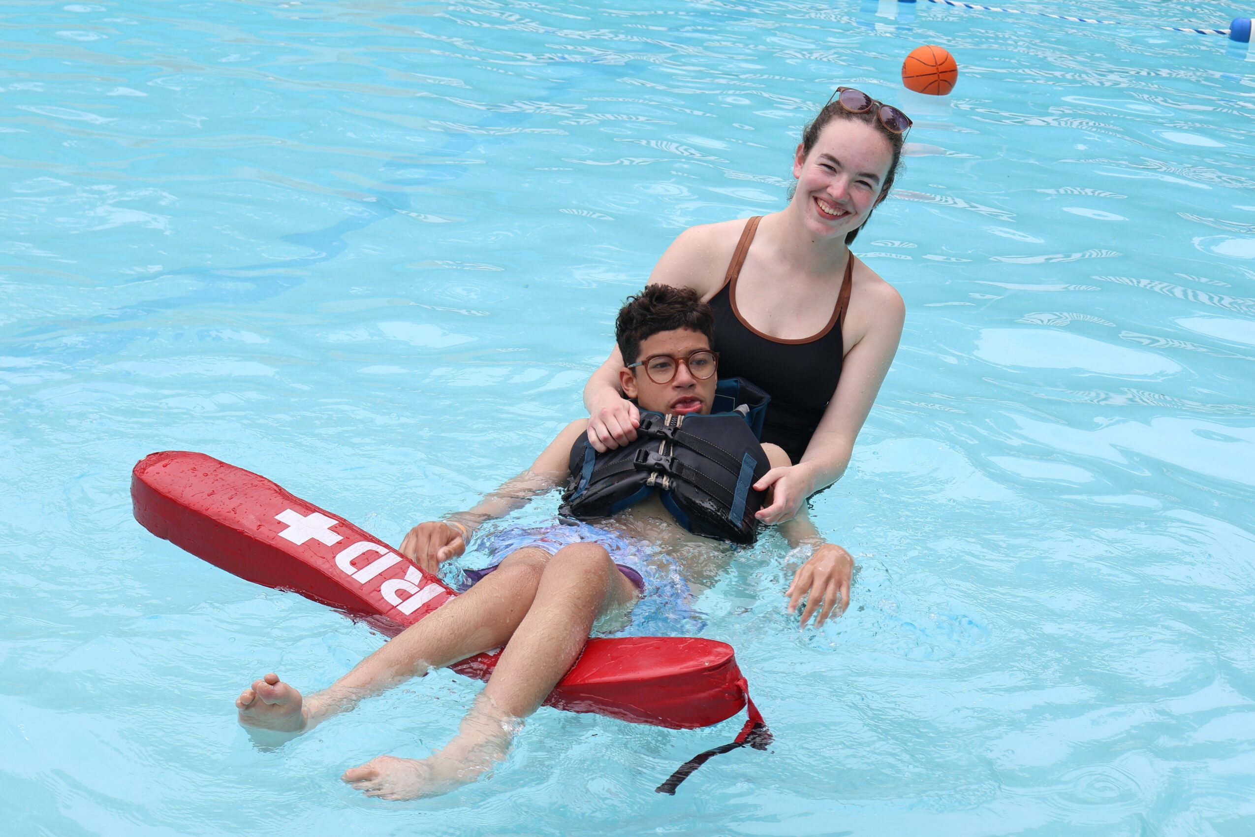 Woman and child in a swimming pool, woman holding the child on a red float, both smiling, water around them.