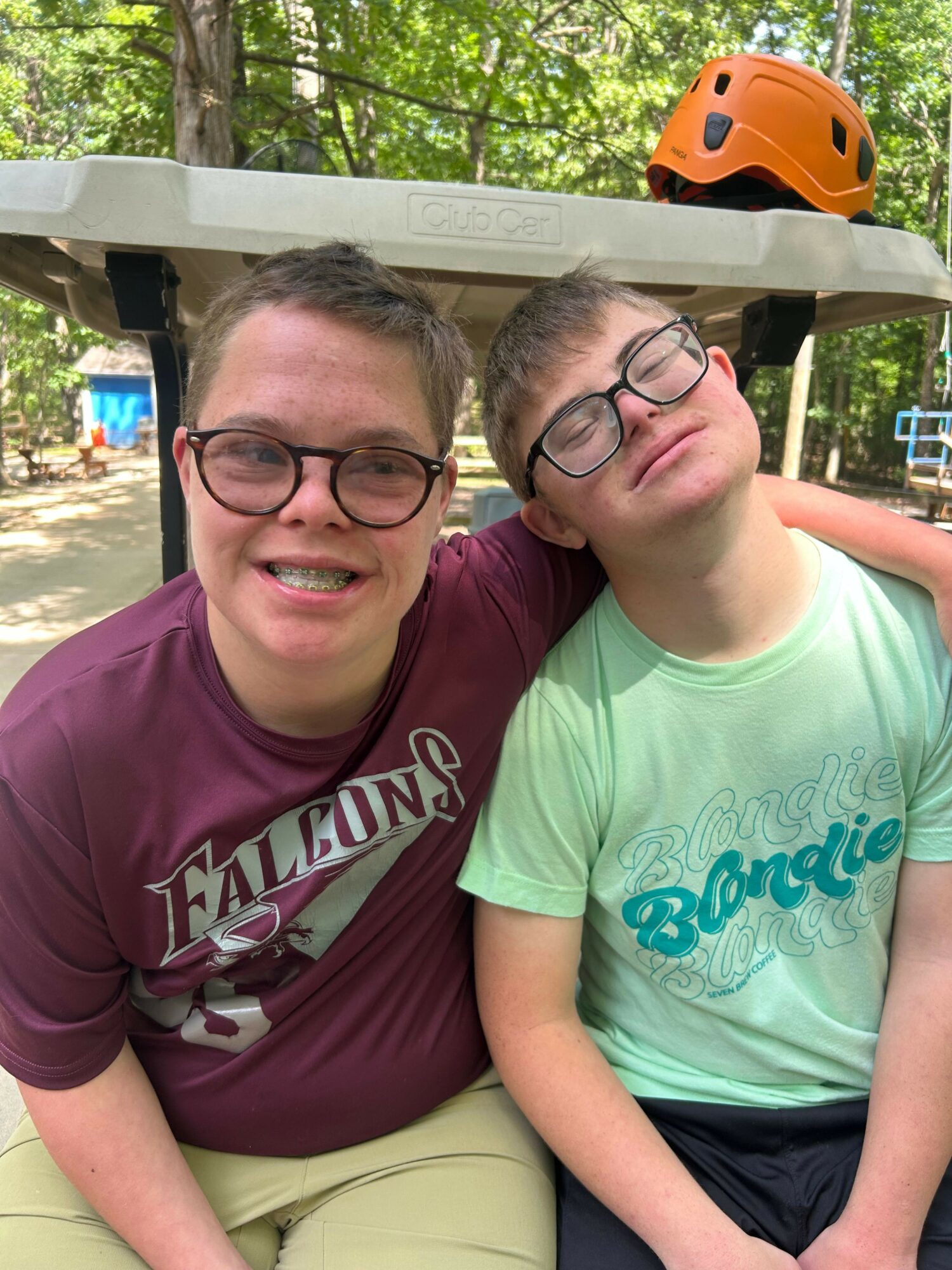 Two people with glasses smiling outdoors, one wearing a green shirt and the other a maroon shirt, with trees in background.