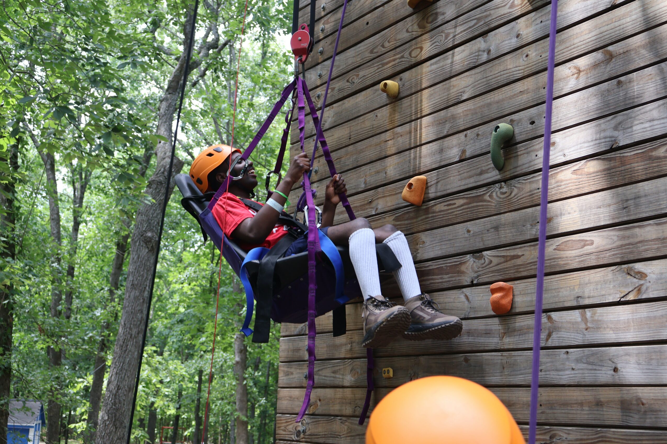 Child in harness and helmet sitting on a swing attached to a wooden climbing wall outdoors, surrounded by trees.