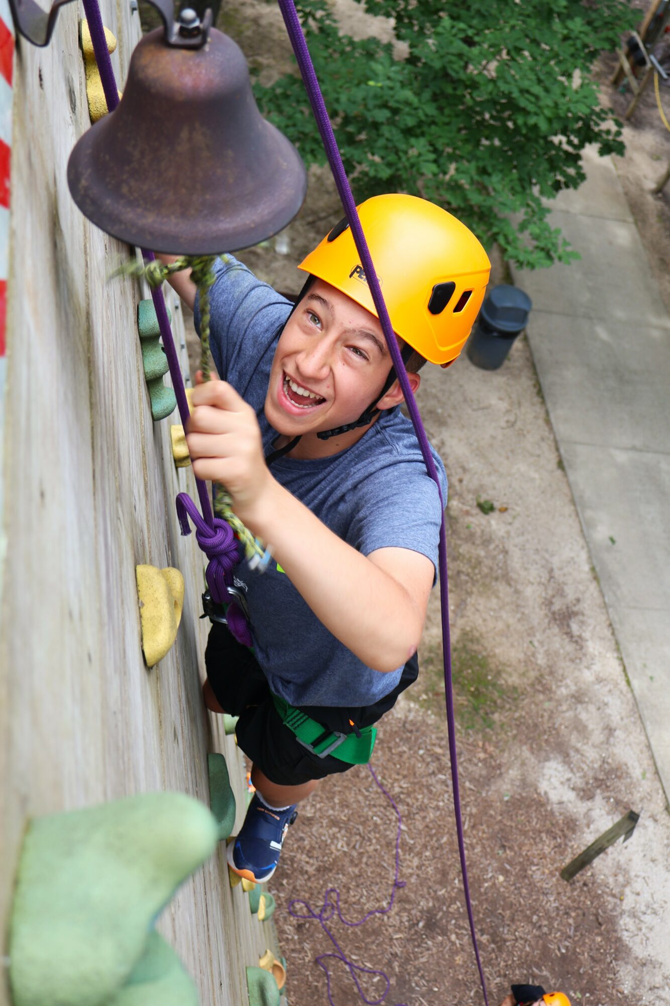 Young person wearing a yellow helmet climbing a wooden wall with a bell, smiling, outdoors.